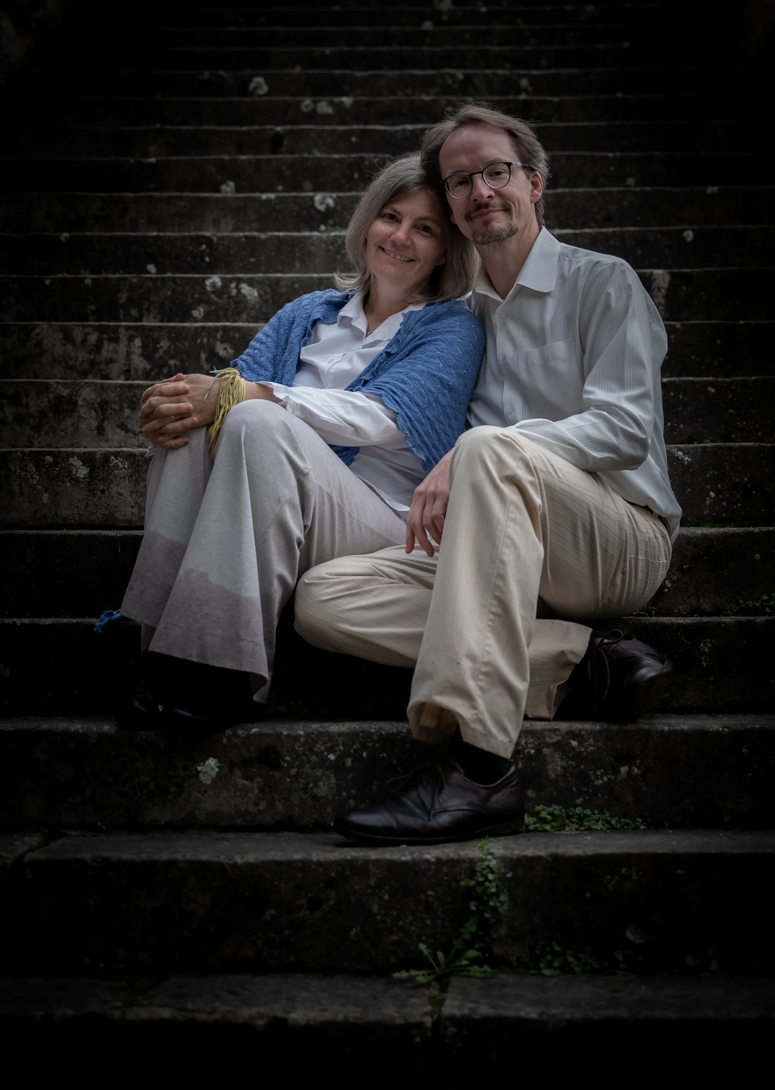 Ein älteres Paar sitzt auf einer Treppe im Freien, beide lachen und wirken glücklich. Der Mann trägt eine weiße Hemd und beige Hosen, die Frau trägt eine weiße Bluse, eine blaue Jacke und eine beige Hose.