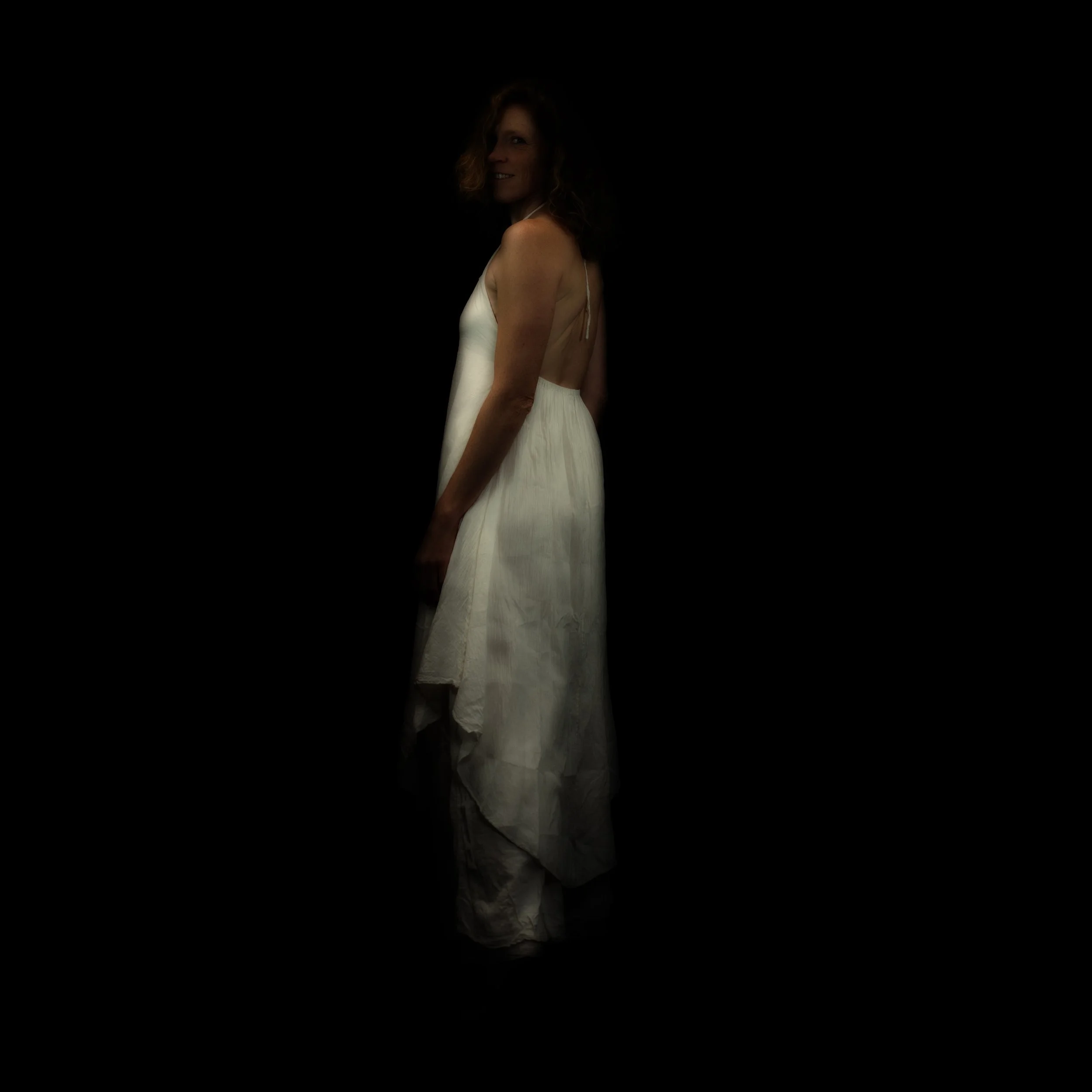 Frau in weißem Kleid steht vor schwarzem Hintergrund, nur halb im Licht, lächelt leicht.