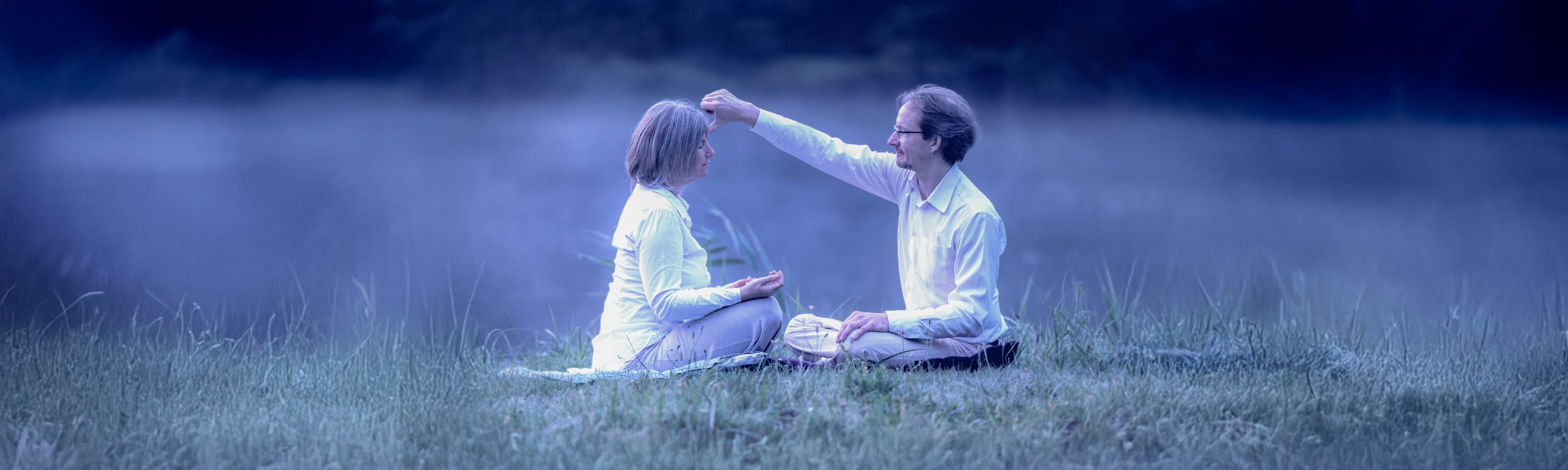 Ein Mann tröstet eine Frau, beide sitzen auf einer Wiese bei Dämmerung.