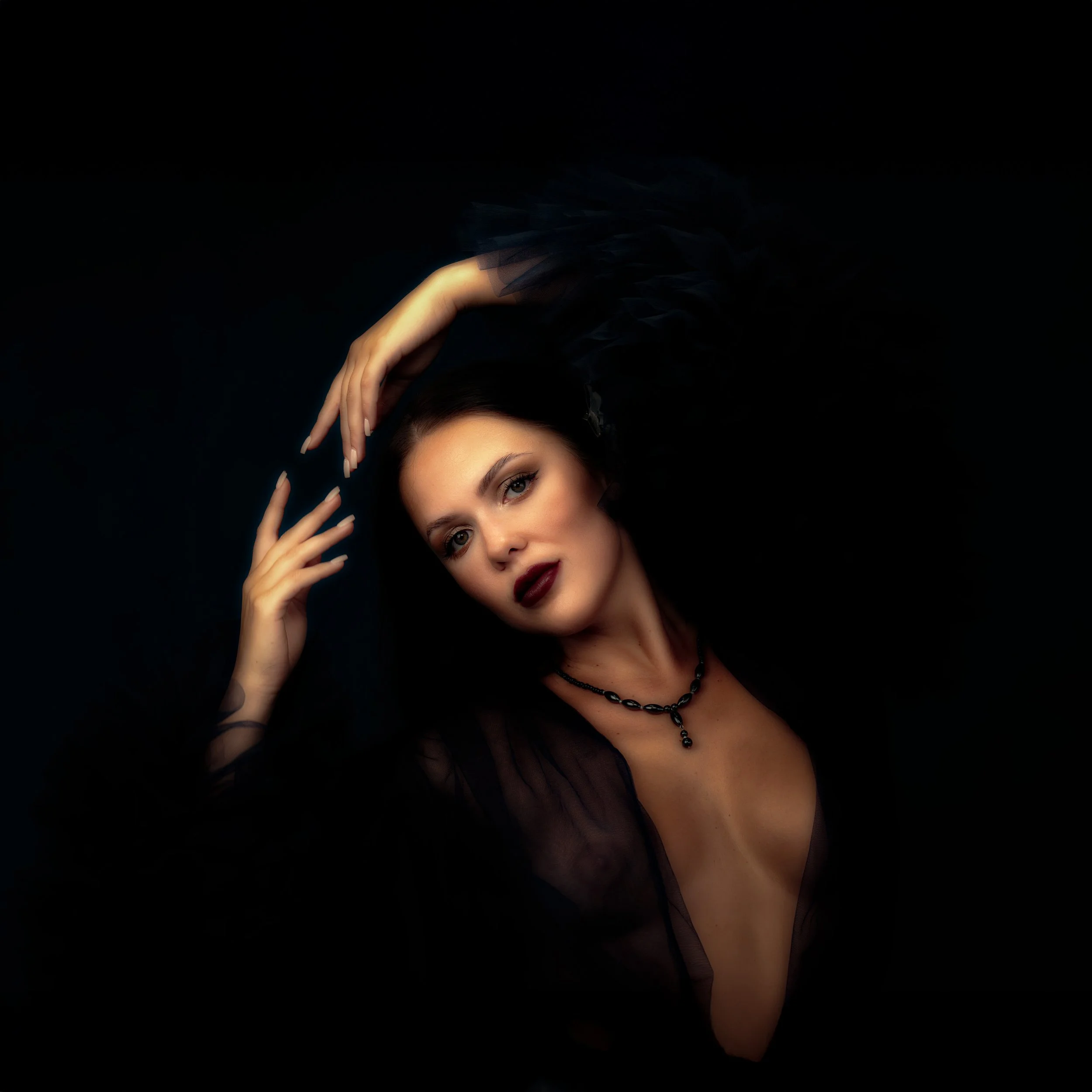 Porträt einer Frau mit dunklem Make-up und schwarzen Kleid vor schwarzem Hintergrund, die ihr Haar stylt.