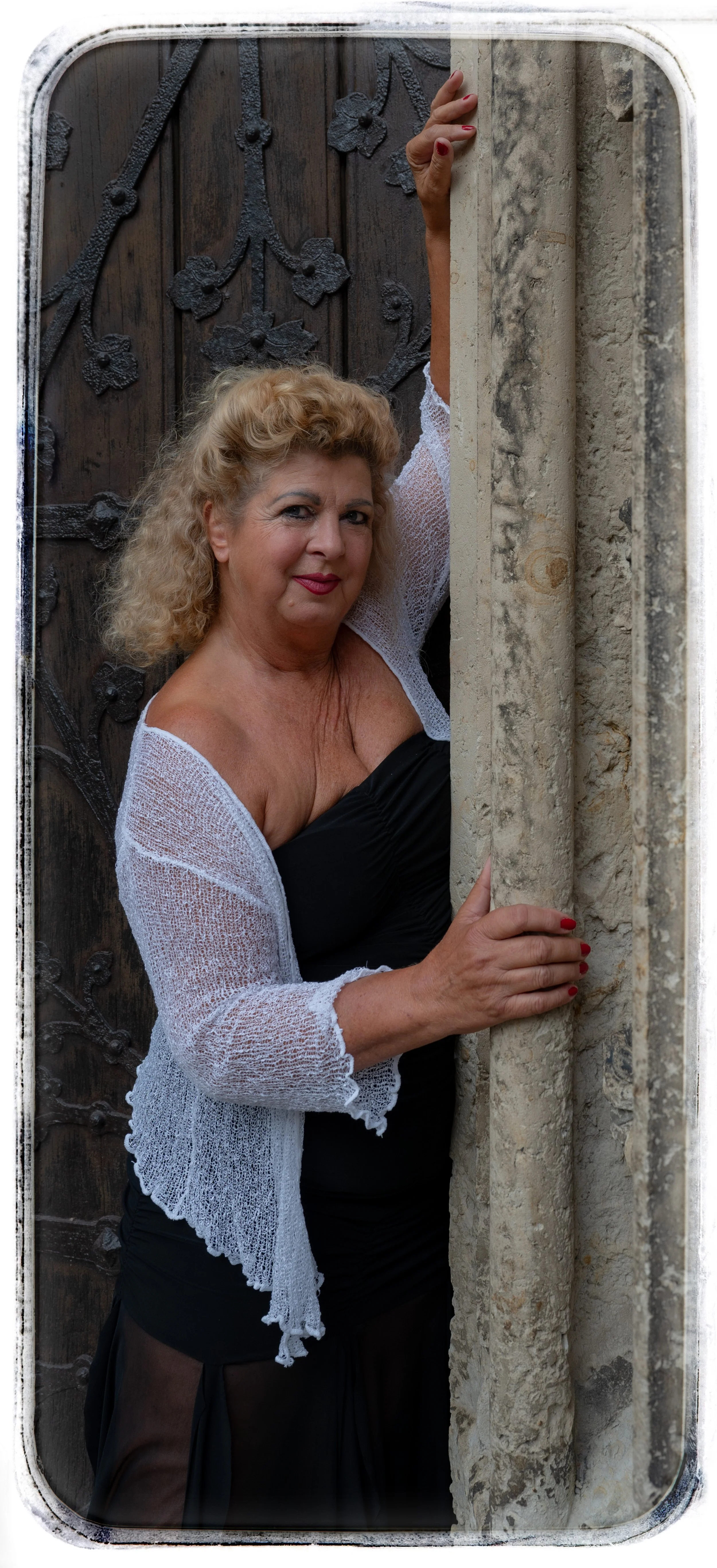 Eine ältere Frau mit lockigem, blondem Haar, trägt ein schwarzes Kleid und eine weiße, lockere Jacke, während sie hinter einer Steinmauer und einer Holz-Tür hervorschaut.