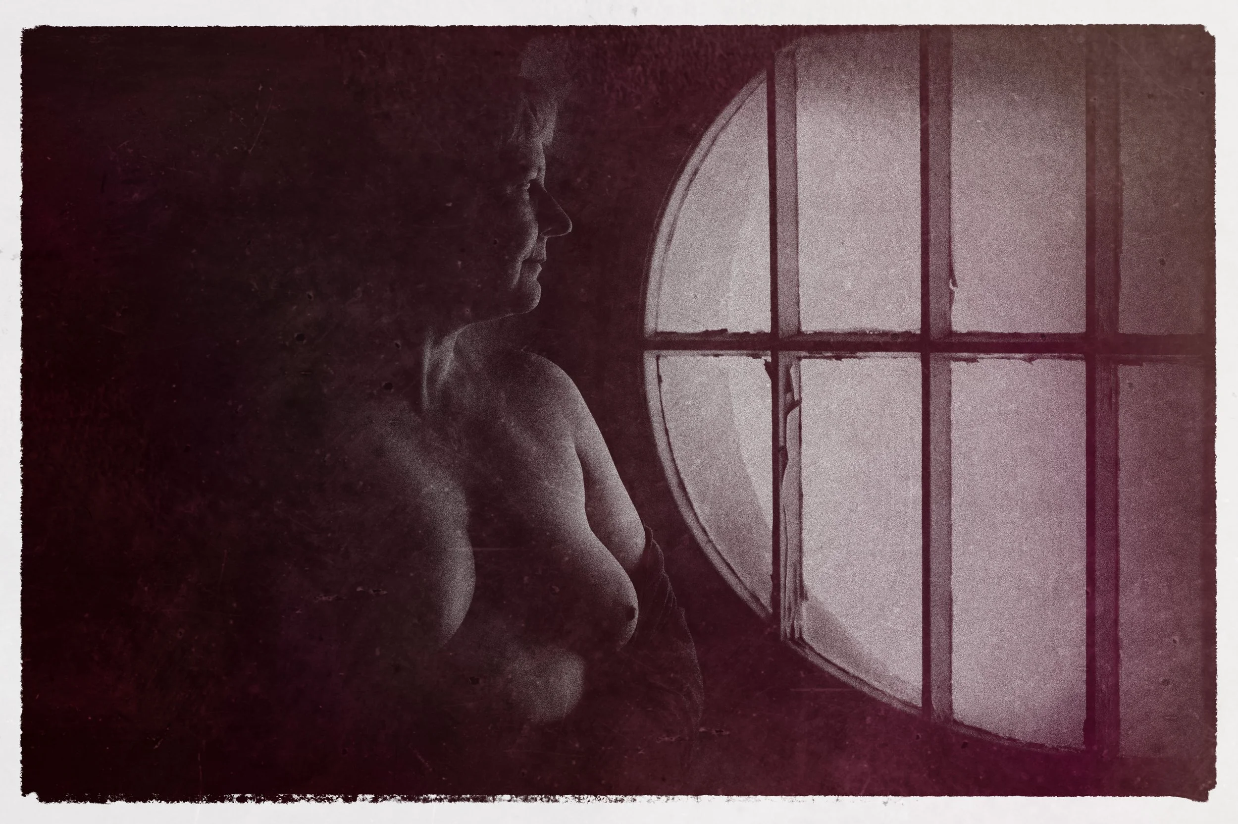 Eine nackte Frau schaut nach draußen durch ein rundes Fenster, im Dunkeln.