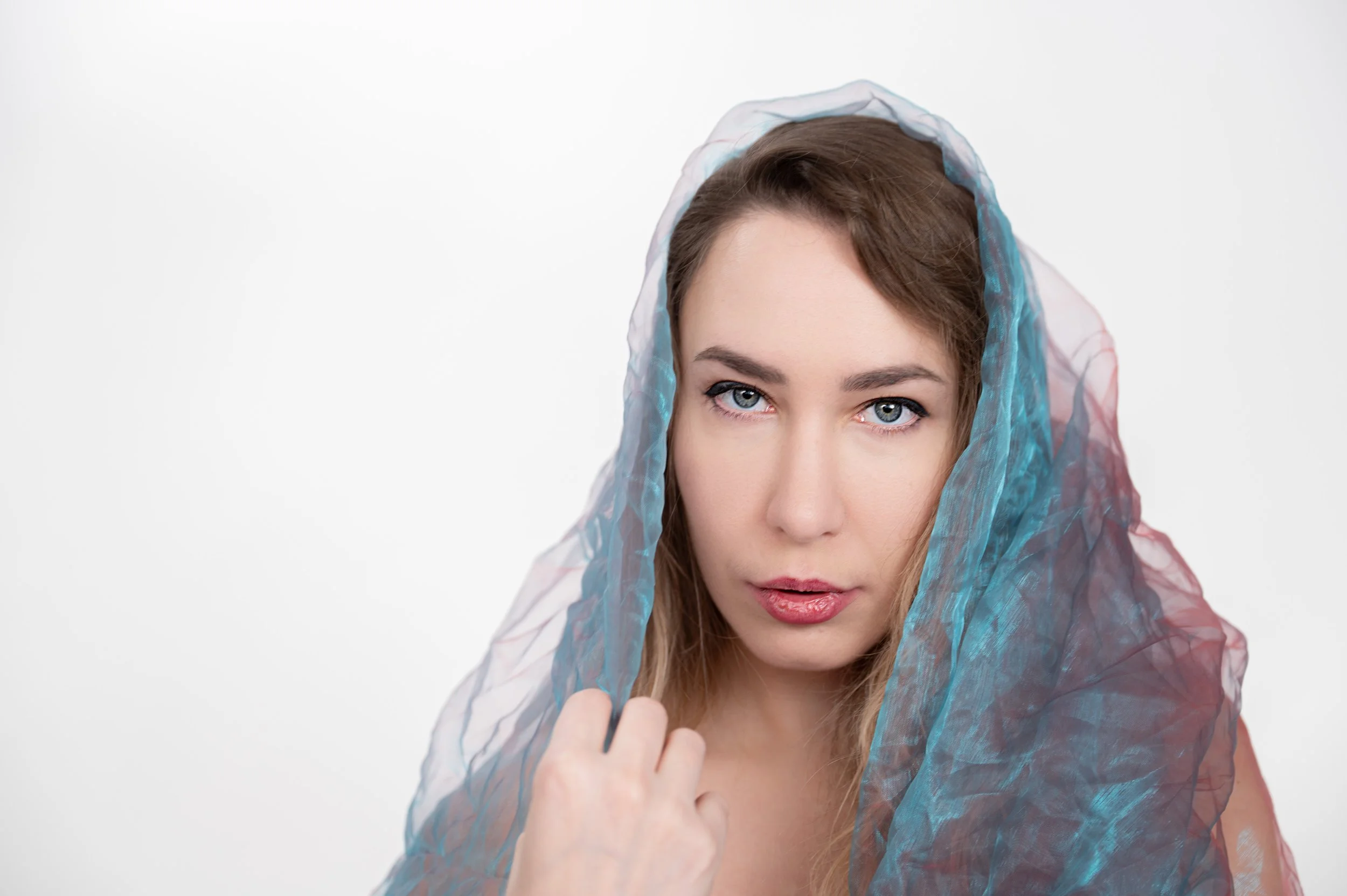 Junge Frau mit blauer und roter durchsichtiger Tüll über ihrem Kopf, Blick in die Kamera, neutraler weißer Hintergrund.