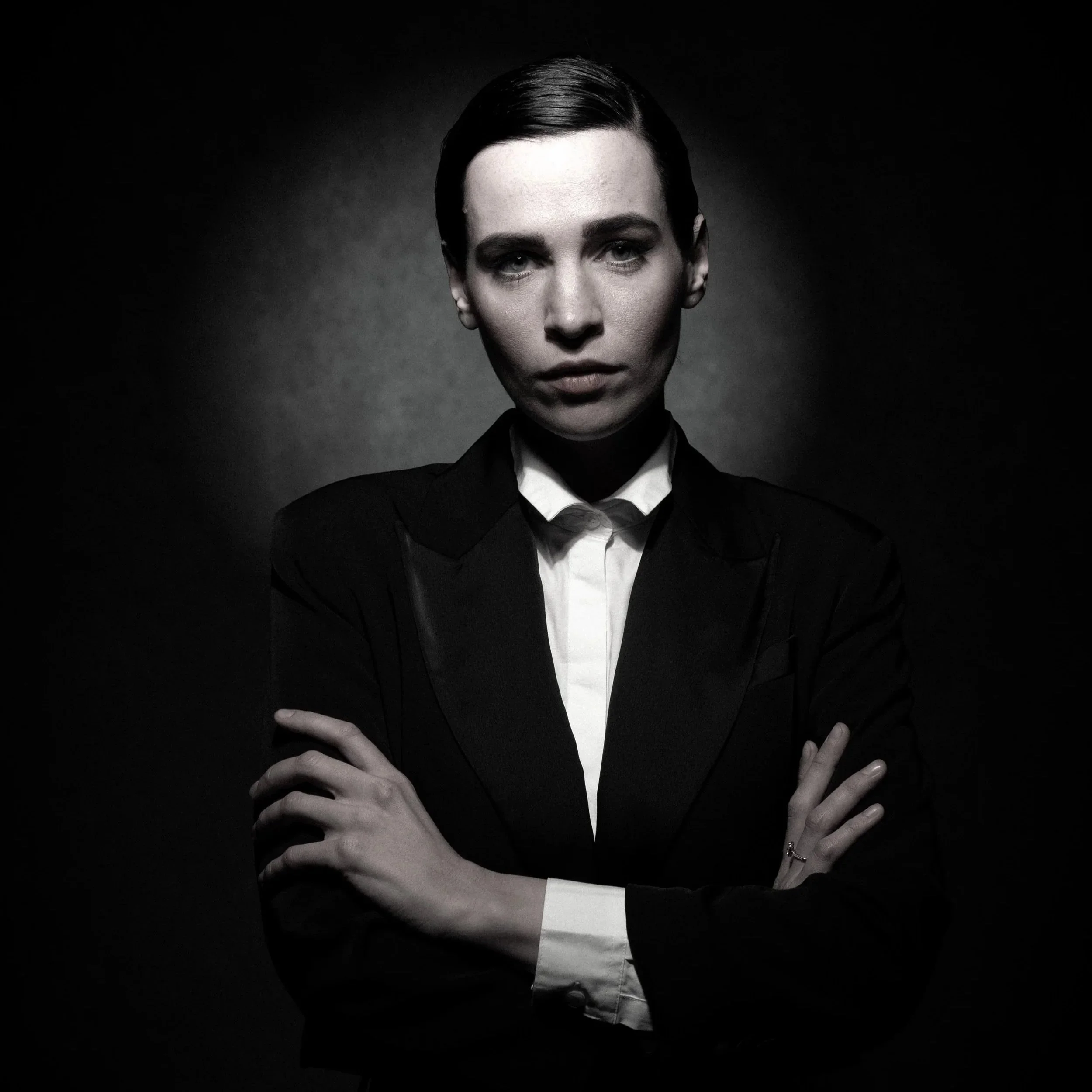 Schwarz-Weiß-Foto einer ernst aussehenden Frau in einem schwarzen Anzug mit hochgesteckten Haaren, Arme verschränkt, Blick direkt in die Kamera.