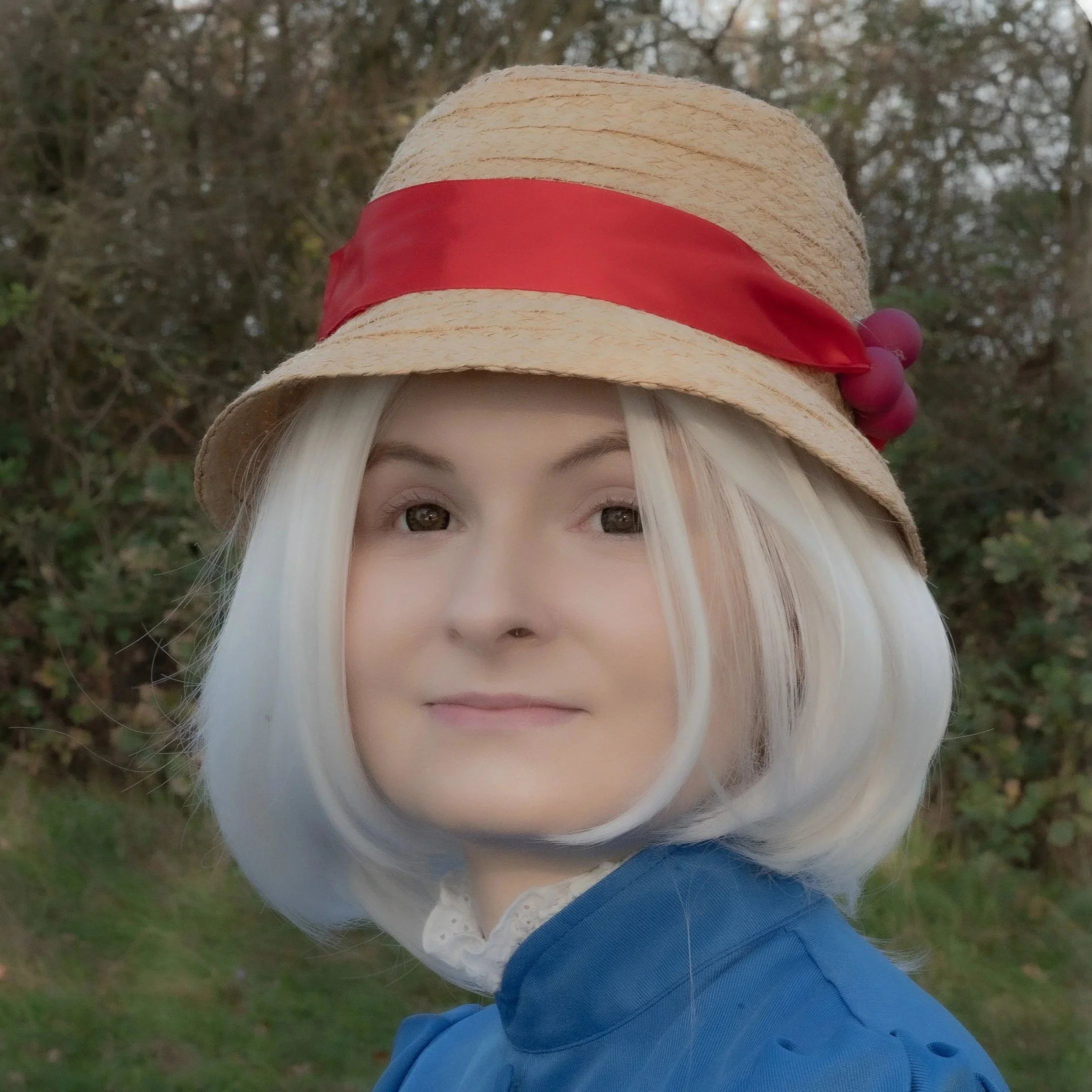 Frau mit weißem Hut mit rotem Band und Fruchtdekoration, blauer Mantel, im Freien mit Büschen im Hintergrund.