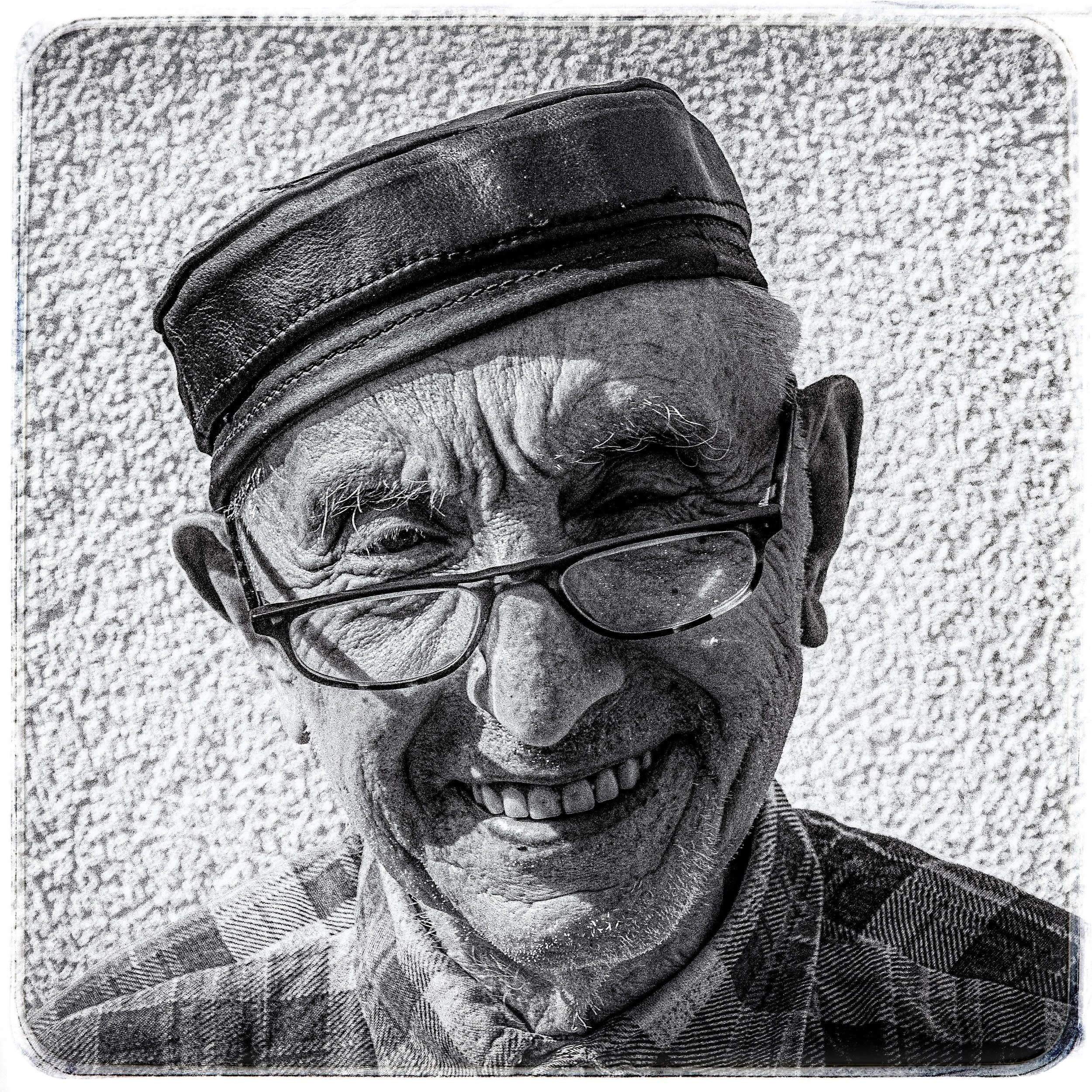 Schwarzweiß-Fotografie eines lachenden älteren Mannes mit Brille, Kappe und kariertem Hemd vor einer textured Wand