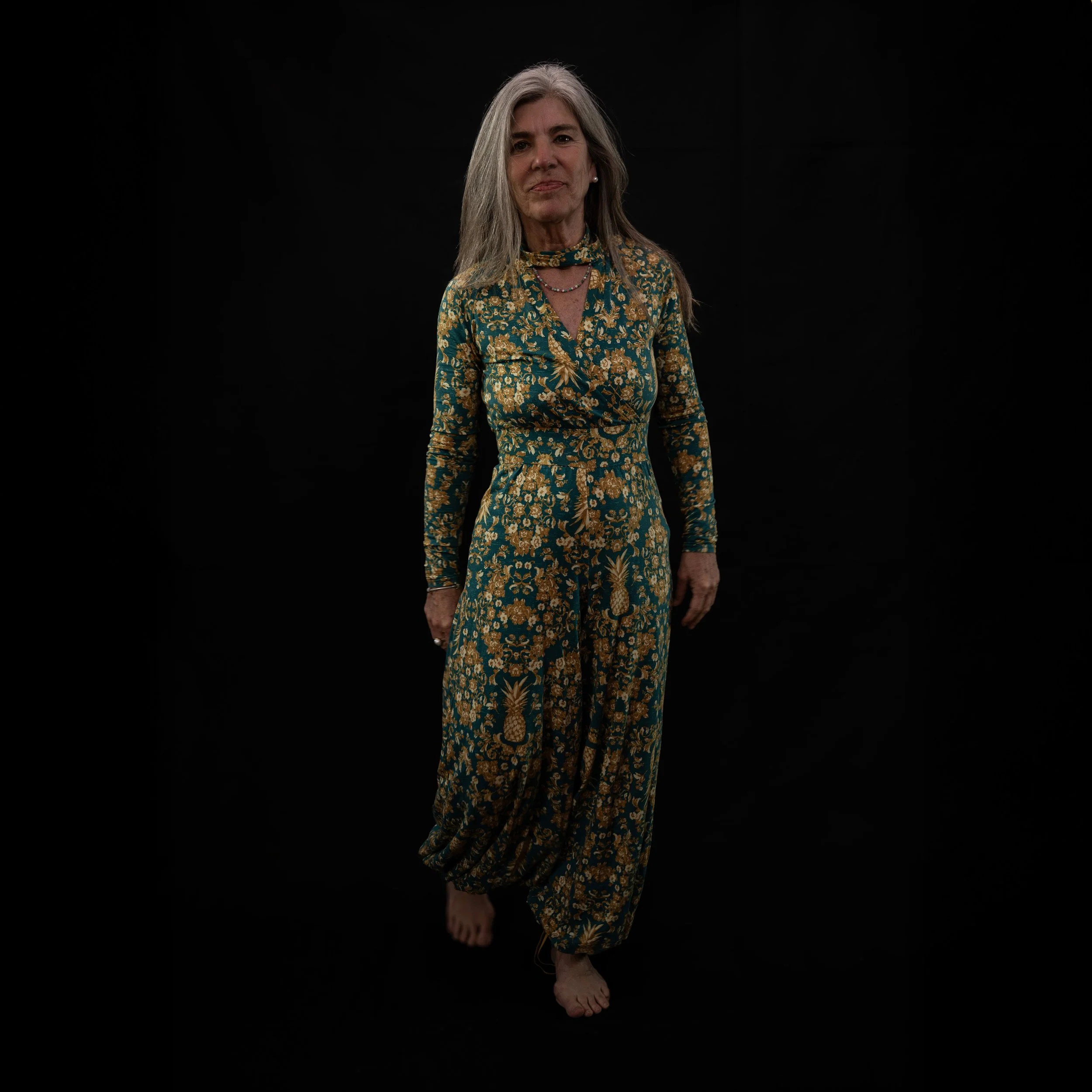 Ältere Frau in gemustertem grünen und goldfarbenem Outfit steht auf schwarzem Hintergrund.