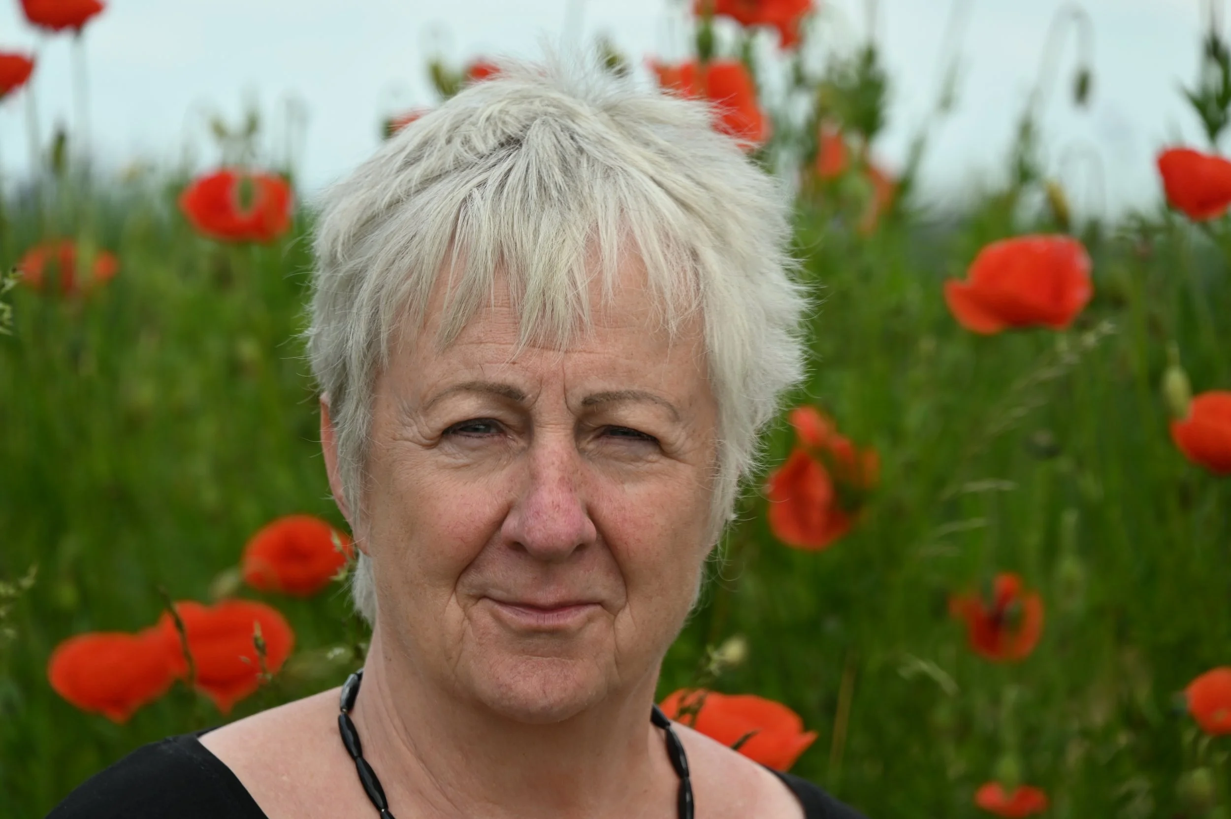 Nahaufnahme einer älteren Frau mit kurzen, weißen Haaren vor einem Feld mit roten Mohnblumen.