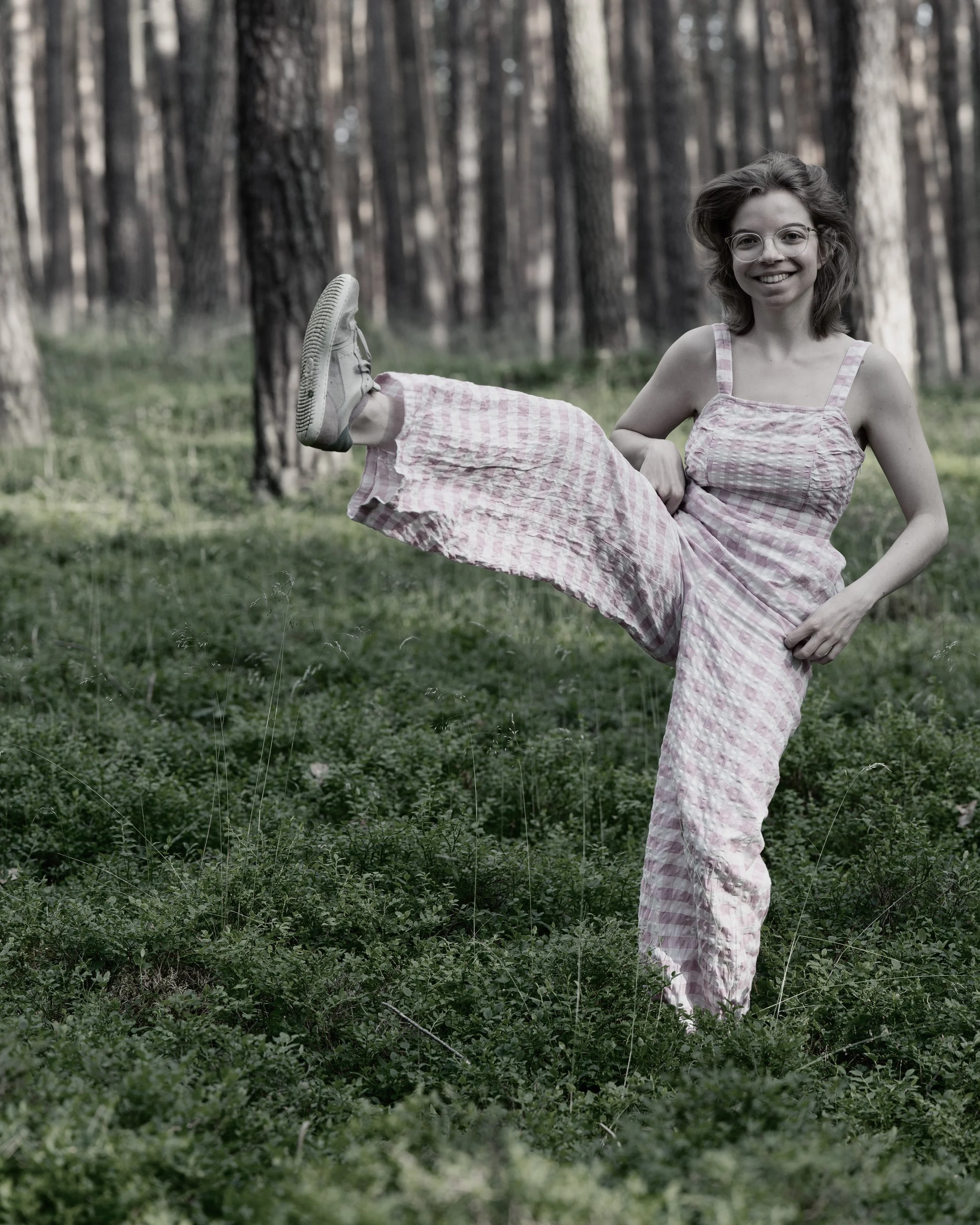 Eine lachende junge Frau in einem Wald, die einen hohen Beinheben-Tanz macht.