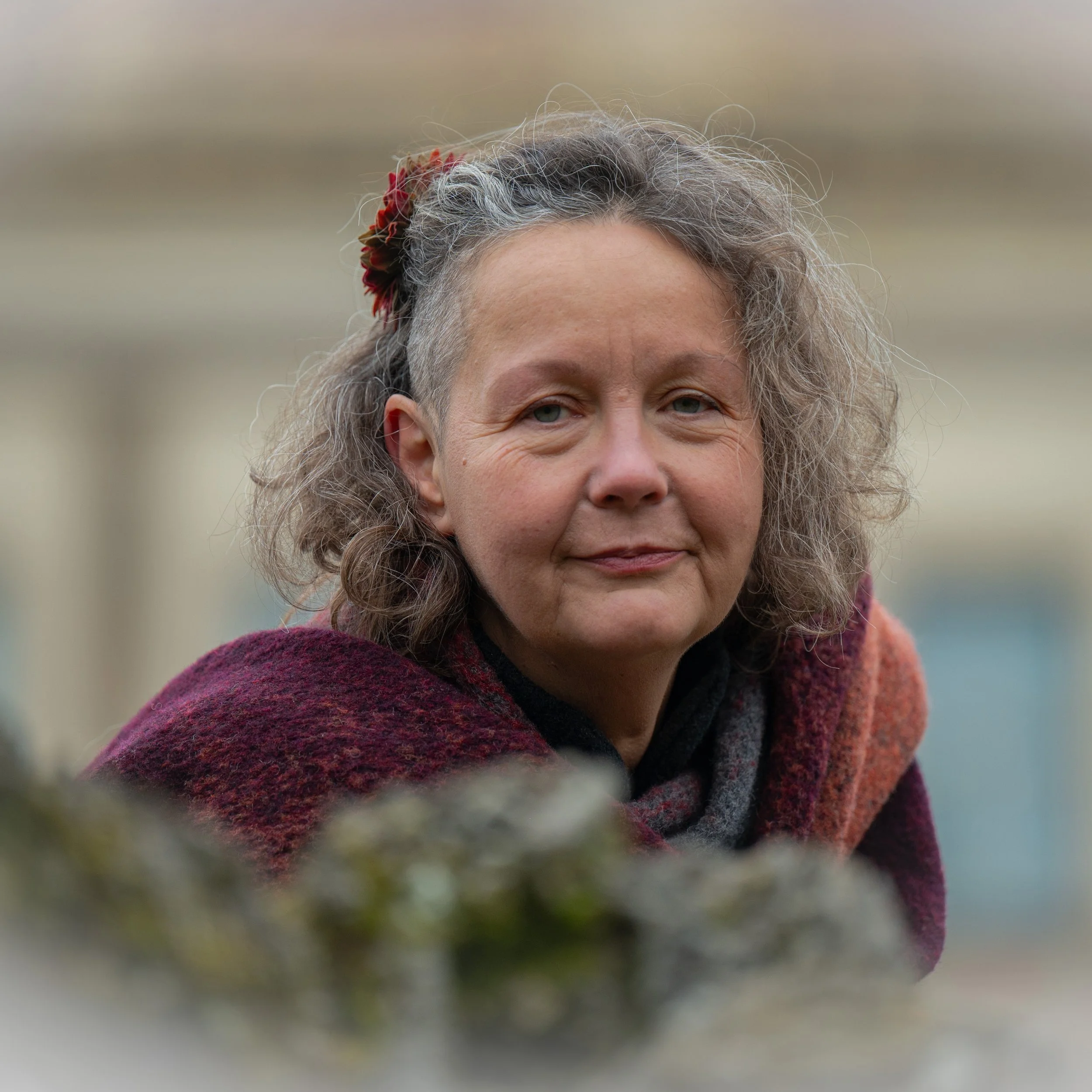 Nahaufnahme einer älteren Frau mit lockigem, grauem Haar, die eine rot-braune Wolljacke trägt, im Hintergrund unscharf.