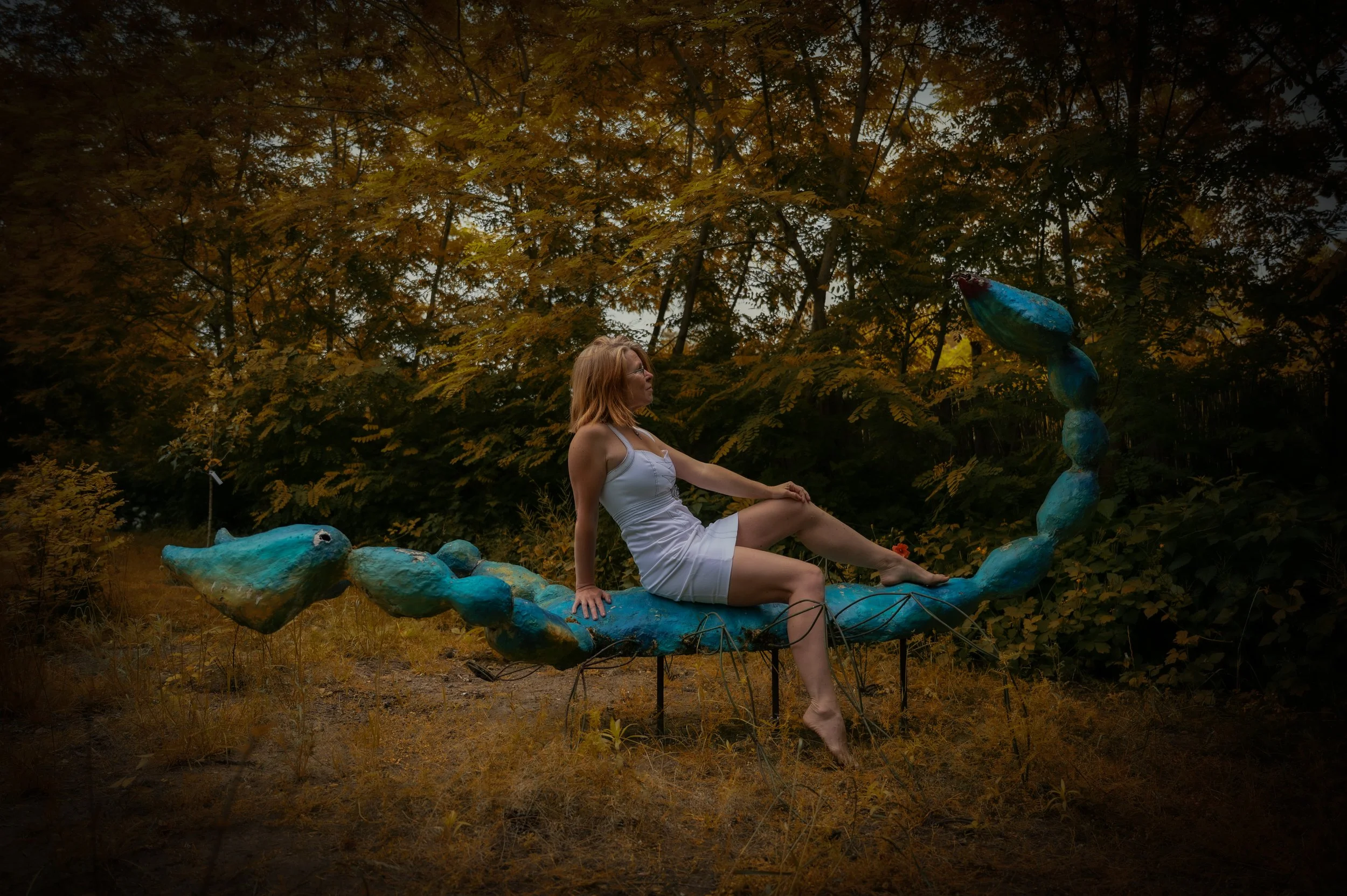 Eine Frau in einem weißen Kleid sitzt auf einer skulpturalen Struktur, die wie eine blaue, geschwungene Kreatur aussieht, in einem Wald mit goldenen Herbstblättern an den Bäumen.