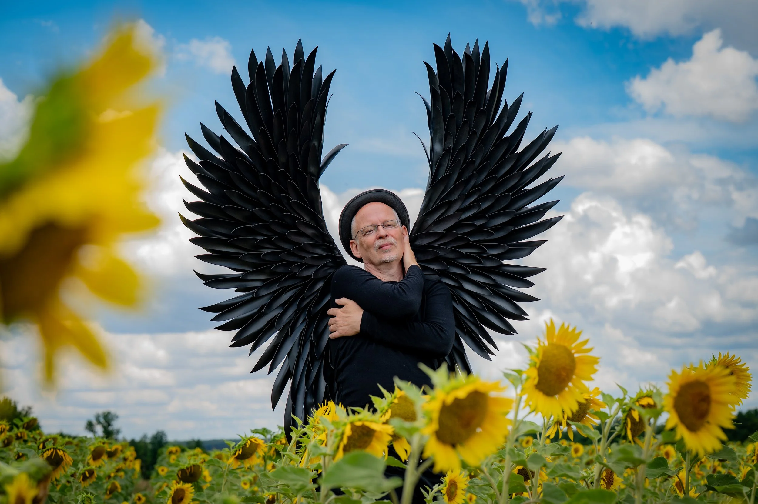 Mensch mit schwarzen Flügeln in einem Sonnenblumenfeld unter einem wolkigen Himmel.