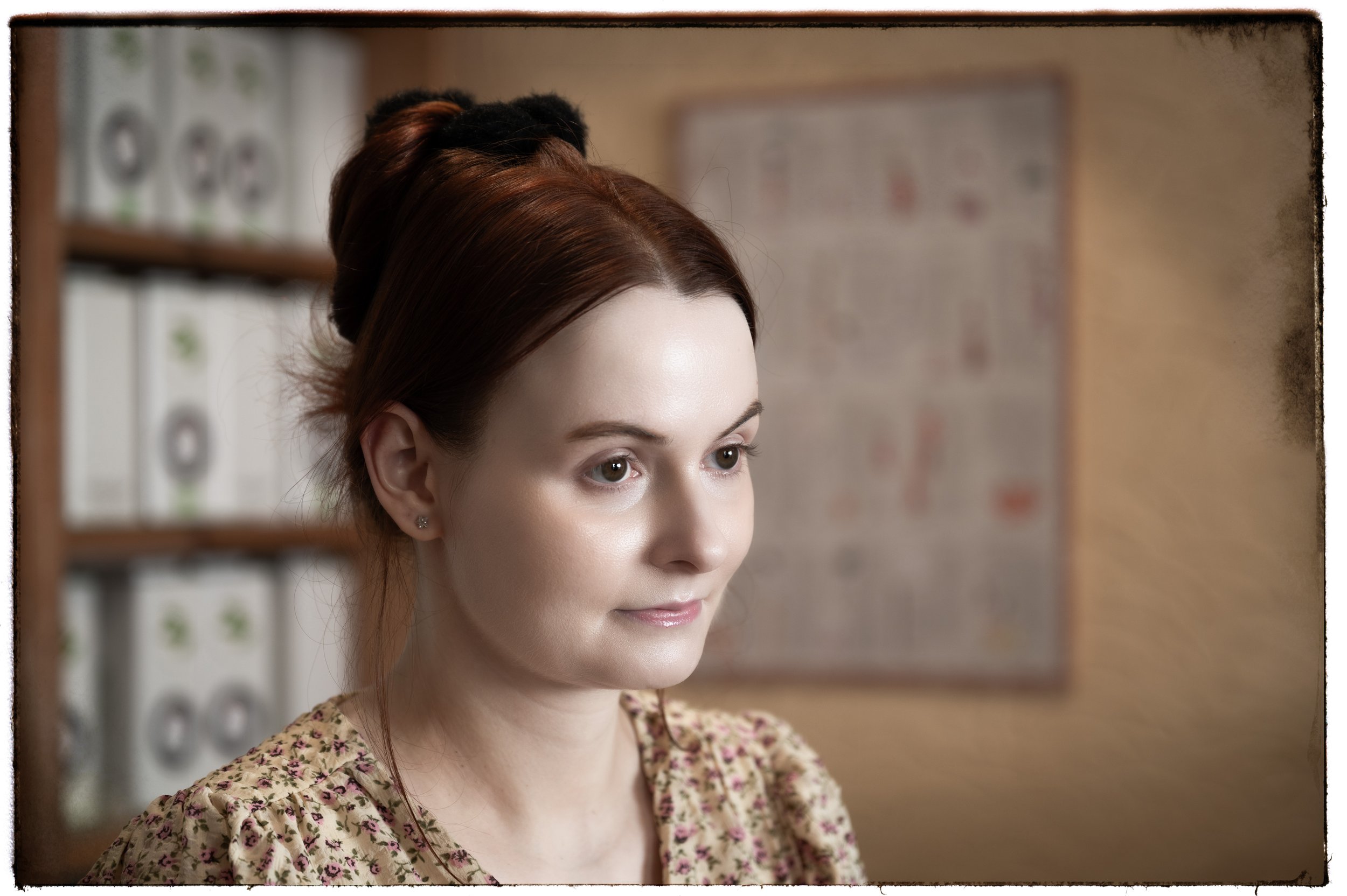 Junge Frau mit braunen Haaren und Blumenmuster-Kleid schaut nach rechts in einem Raum mit Bücherregalen im Hintergrund.