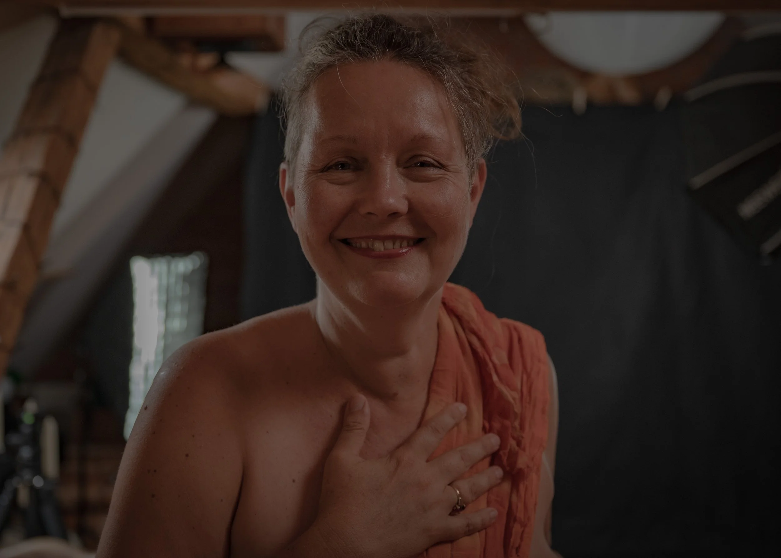 Eine lächelnde Frau mit kurzen grauen Haaren in einem Raum mit Holzbalken, die ein orangefarbenes Kleid trägt, die Hand auf ihrer Brust.