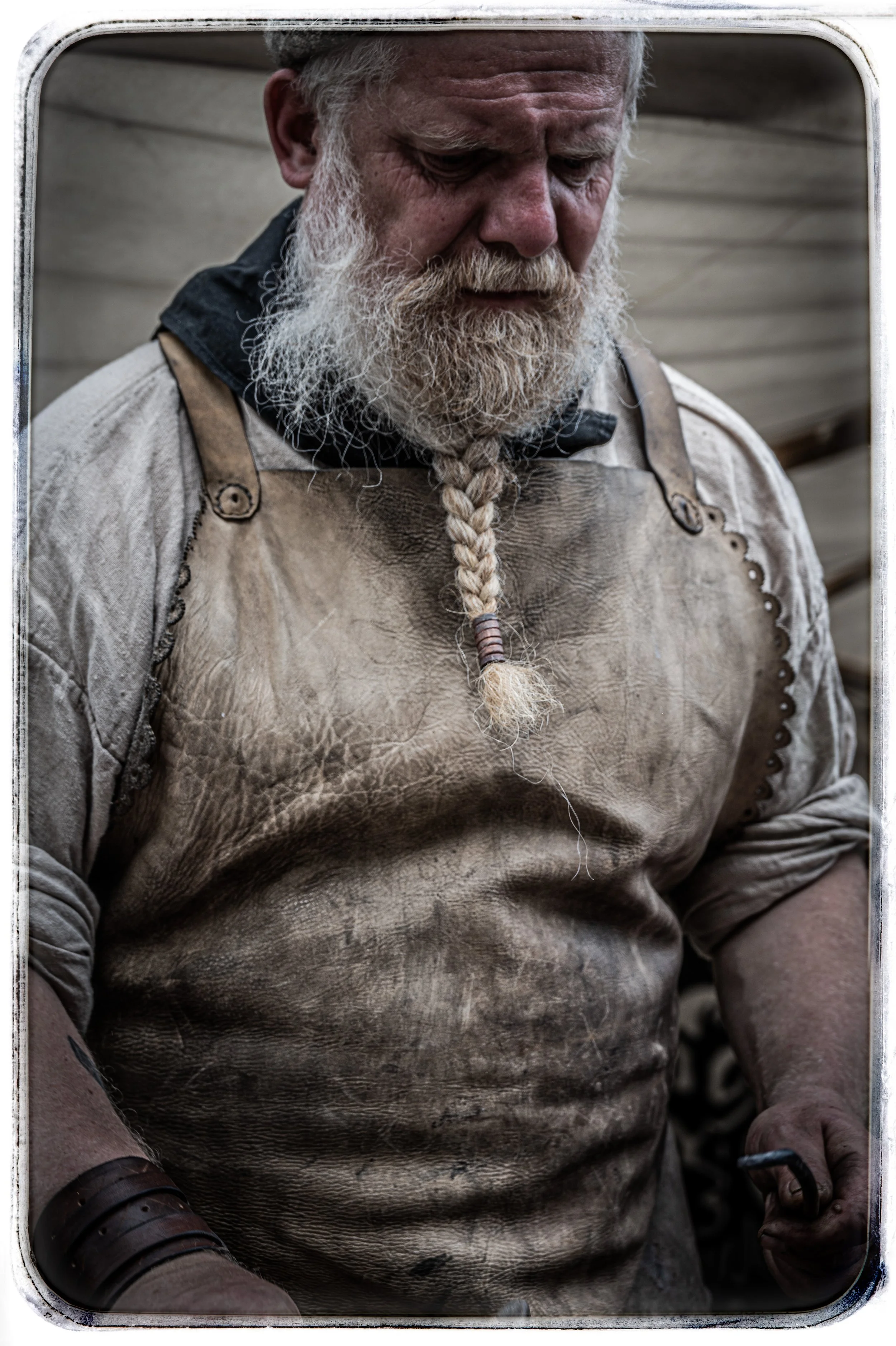 Ein älterer Mann mit langem, weißem Bart und olivgrüner Schürze, die schmutzig ist, steht vor einer Holzwand.