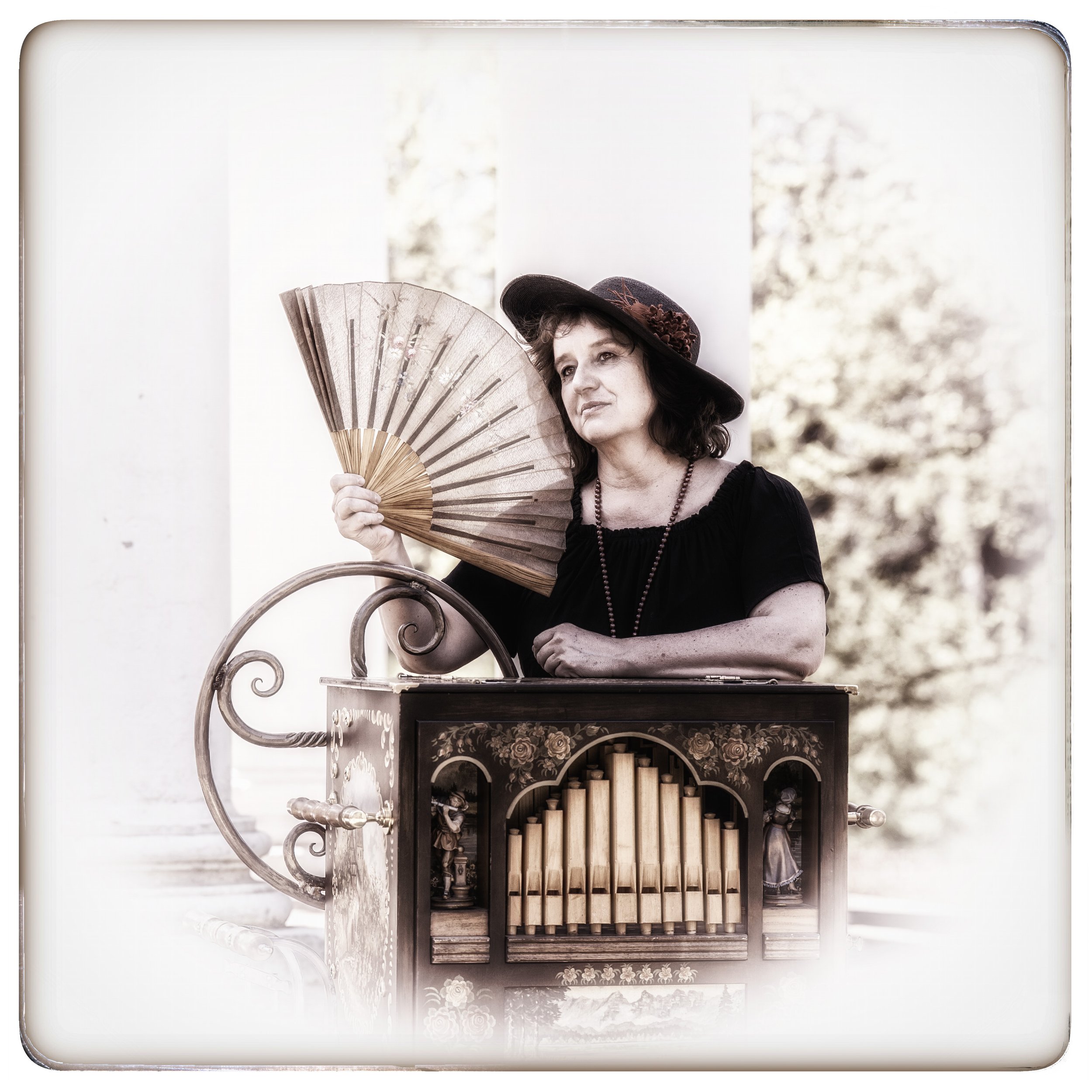 Eine ältere Frau mit dunklem Hut und schwarzem Kleid steht hinter einem alten mechanischen Orgelspiel. Sie hält einen Fächer und blickt nach oben.