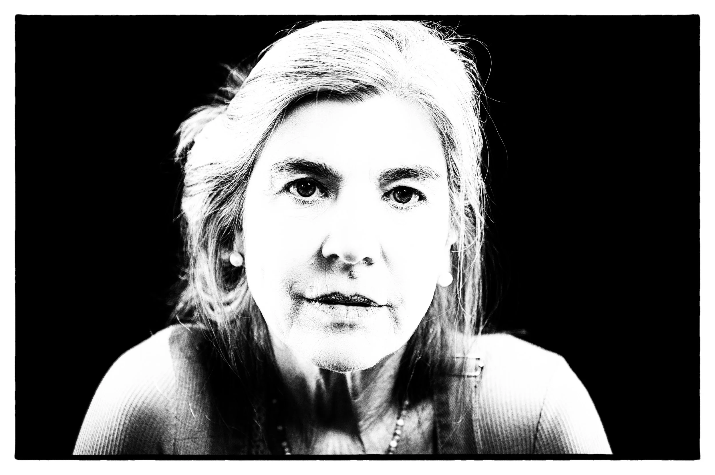 Schwarz-weiß Porträt einer älteren Frau mit langen Haaren, die Lederjacke und Perlen trägt, vor schwarzem Hintergrund.