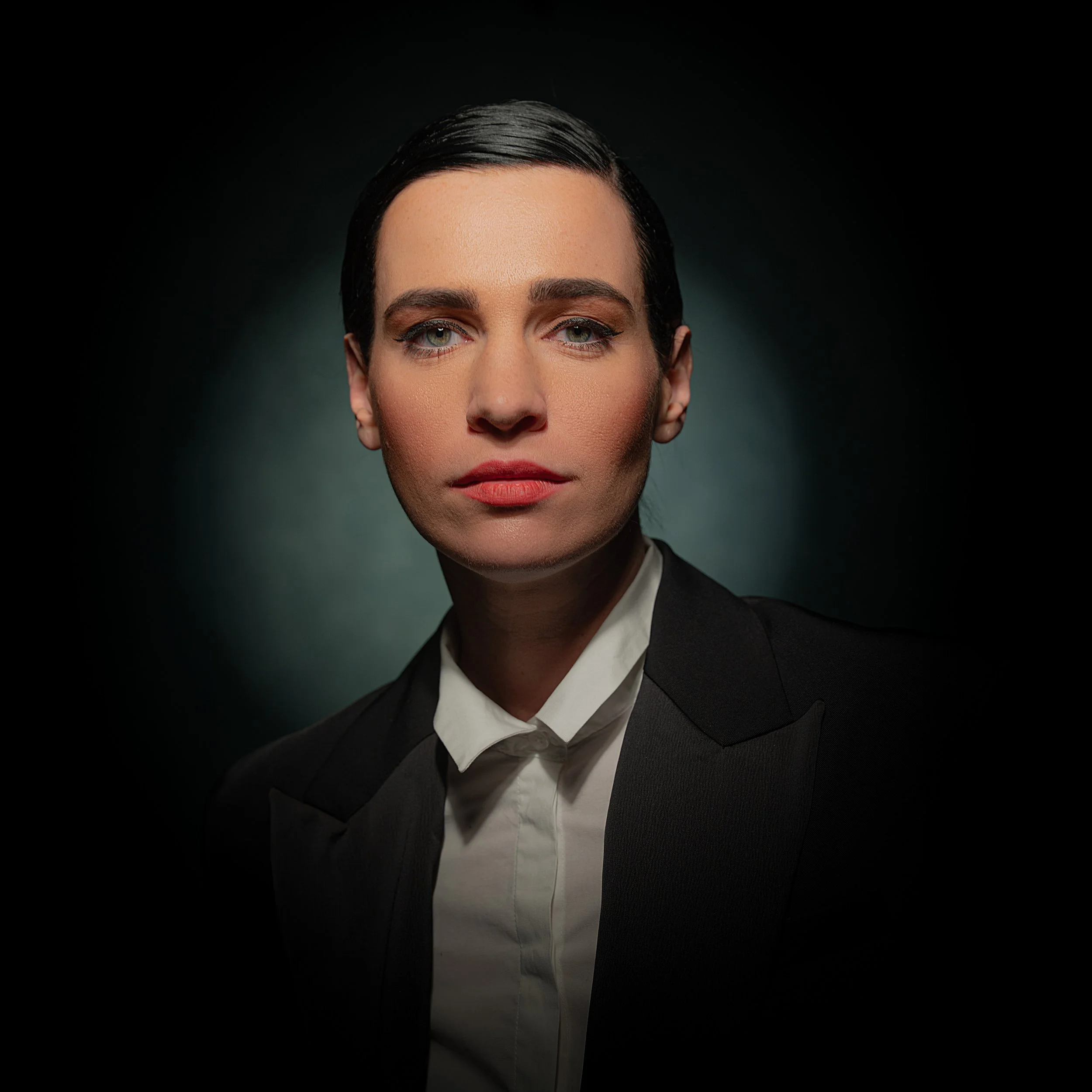 Porträt einer Frau mit dunklem, kurzen Haar, blauer Augen, rotem Lippenstift, in dunkler Kleidung und weißem Hemd, vor dunklem Hintergrund.