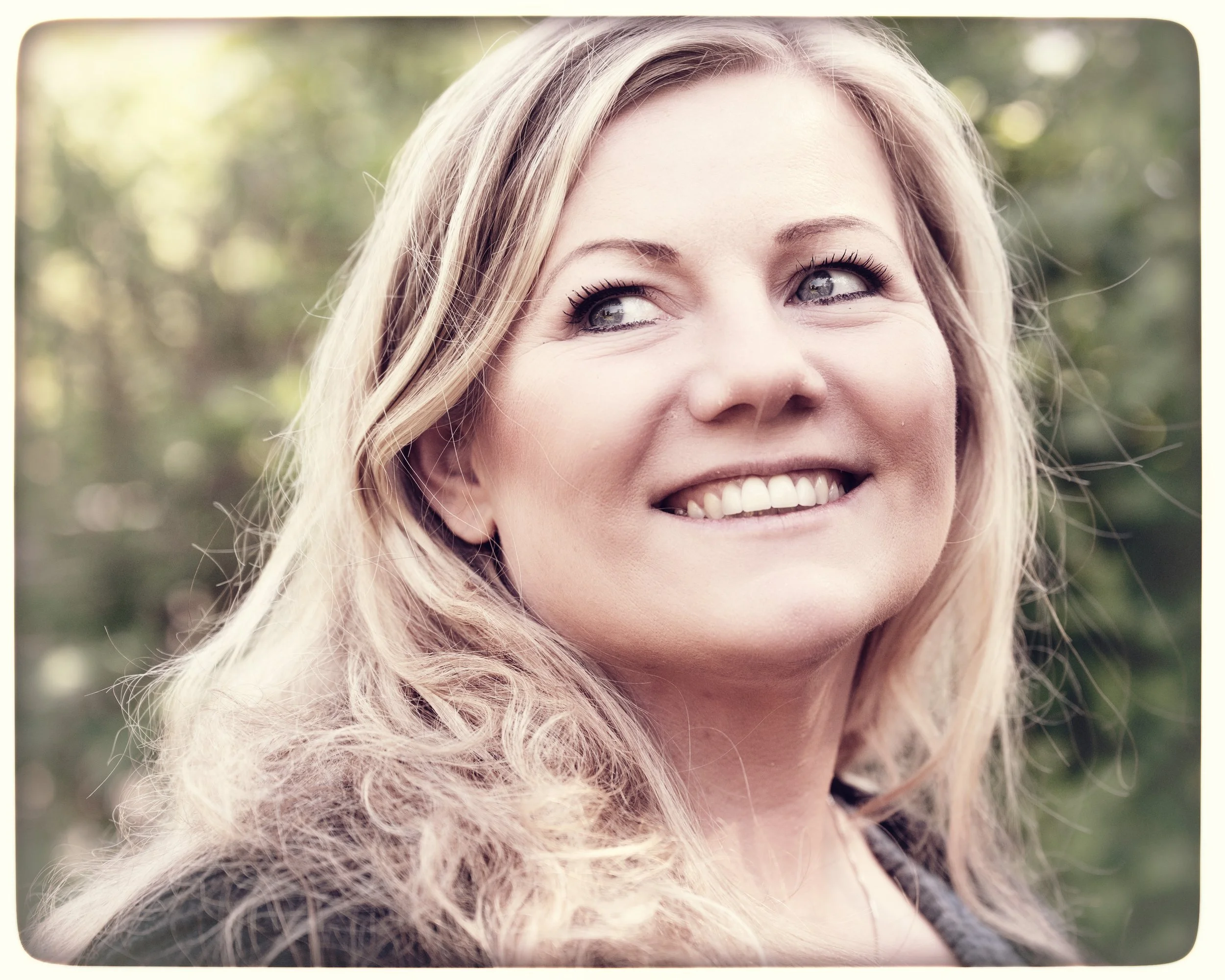 Eine lachende Frau mit langen, lockigen blonden Haaren vor einem natürlichen, grünen Hintergrund.