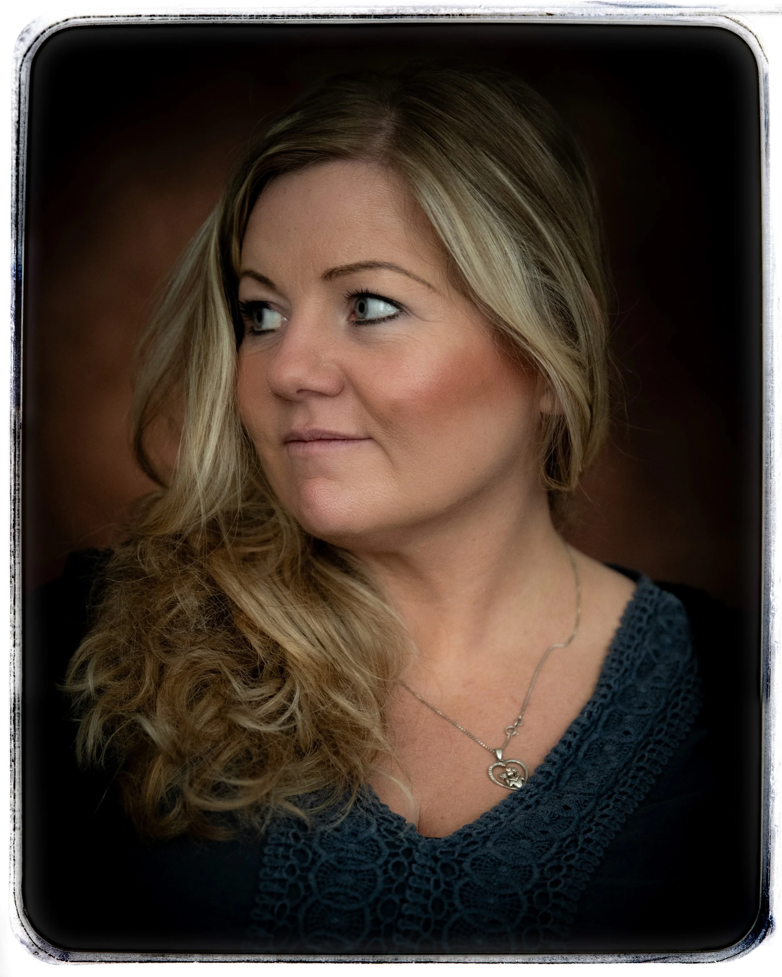 Porträt einer Frau mit lockigem, blonden Haar, die nach rechts schaut, vor dunklem Hintergrund, trägt ein dunkles Oberteil mit Spitze und eine silberne Halskette.