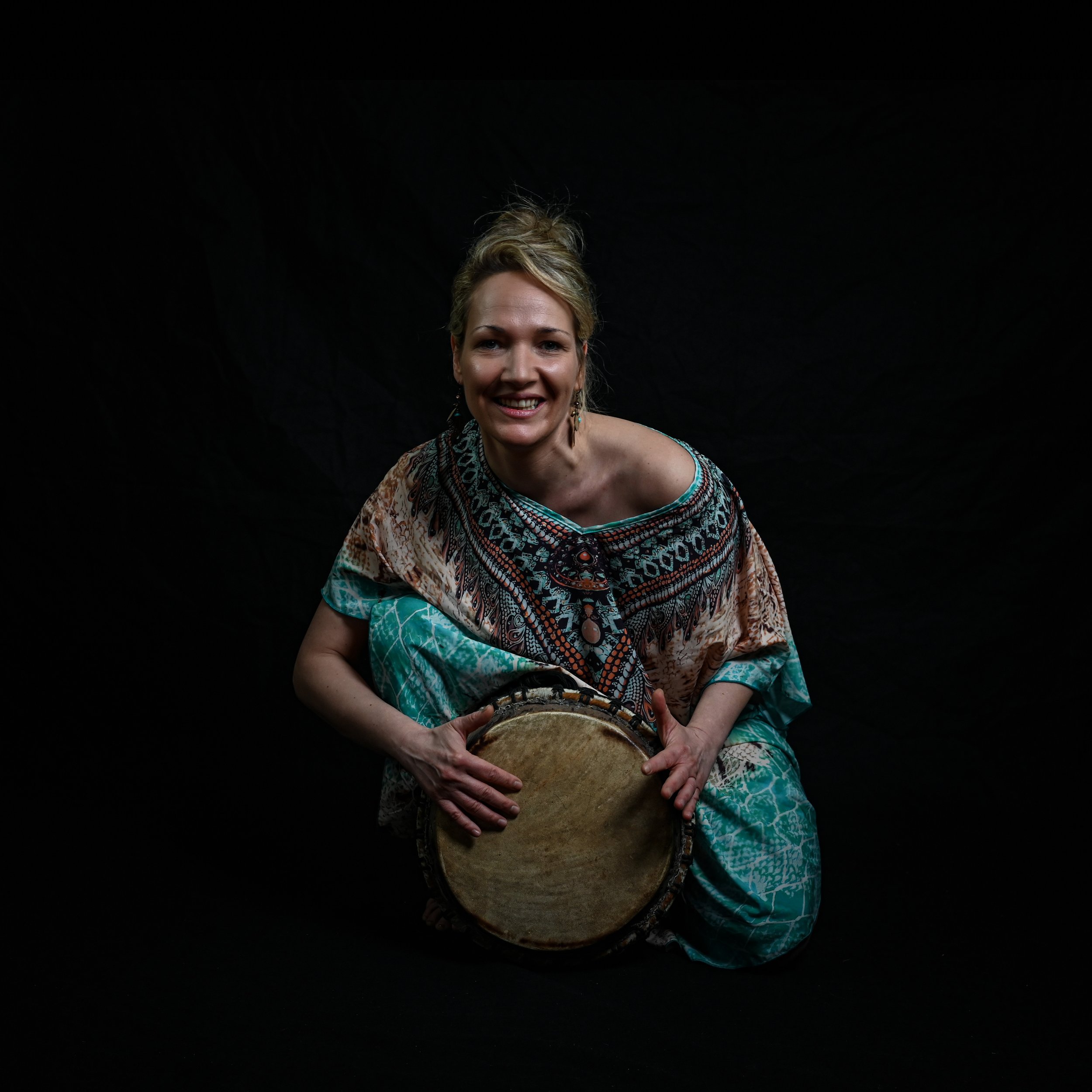 Frau mit buntem Kleid, lachend, hält ein Tamburin gegen einen schwarzen Hintergrund.