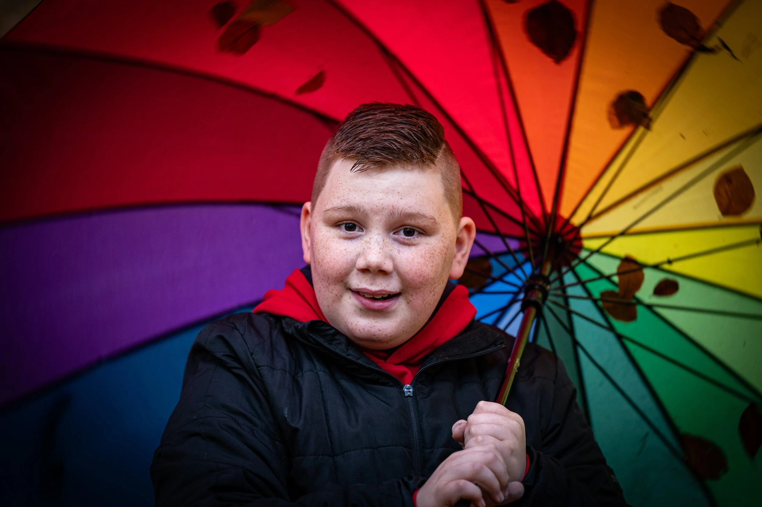 Junger Junge mit roten Haaren und Sommersprossen hält einen bunten Regenschirm vor sich.