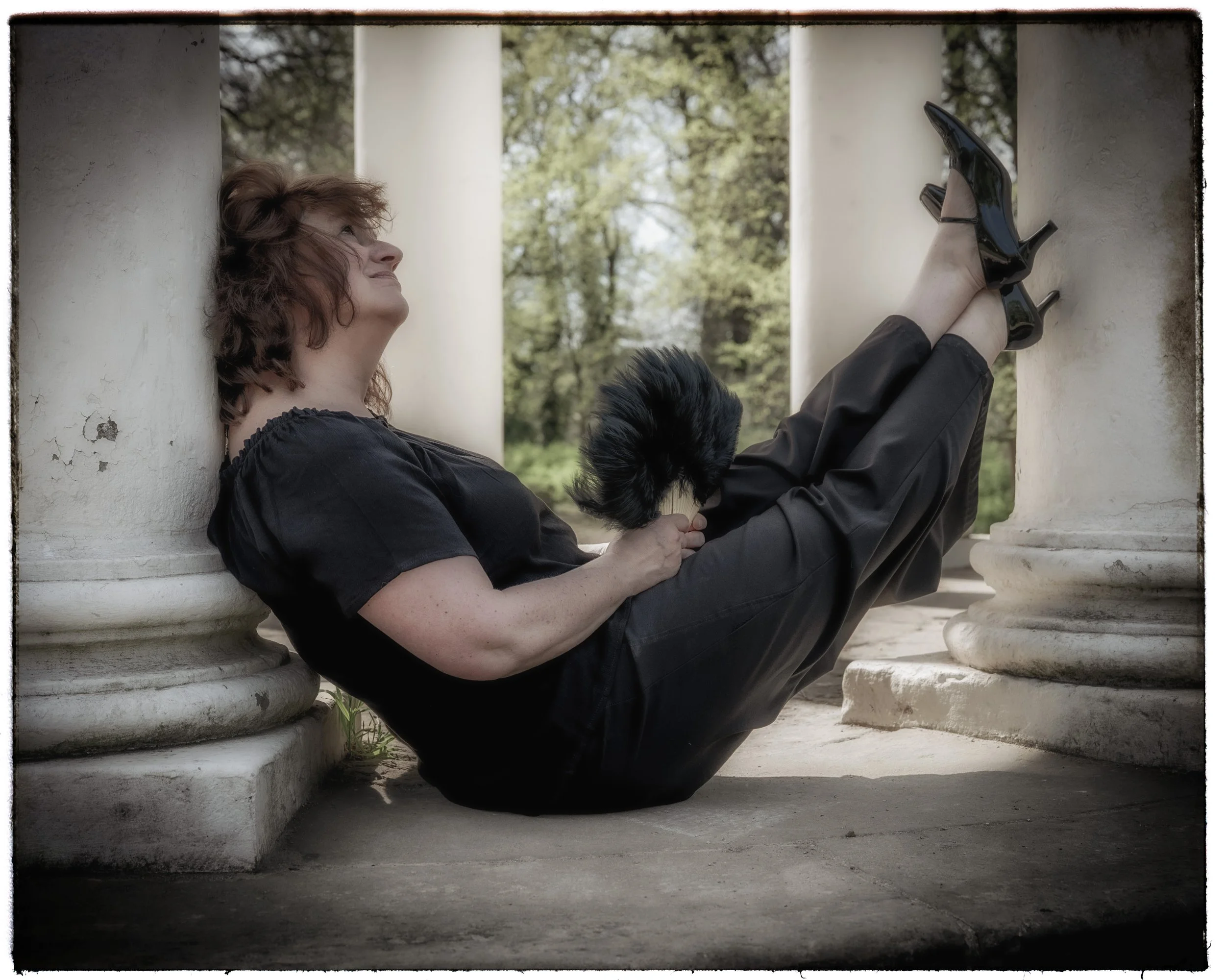 Frau liegt entspannt zwischen zwei Säulen, hält eine schwarze Katze, trägt schwarze Kleidung und High Heels, im Hintergrund Bäume im Freien.