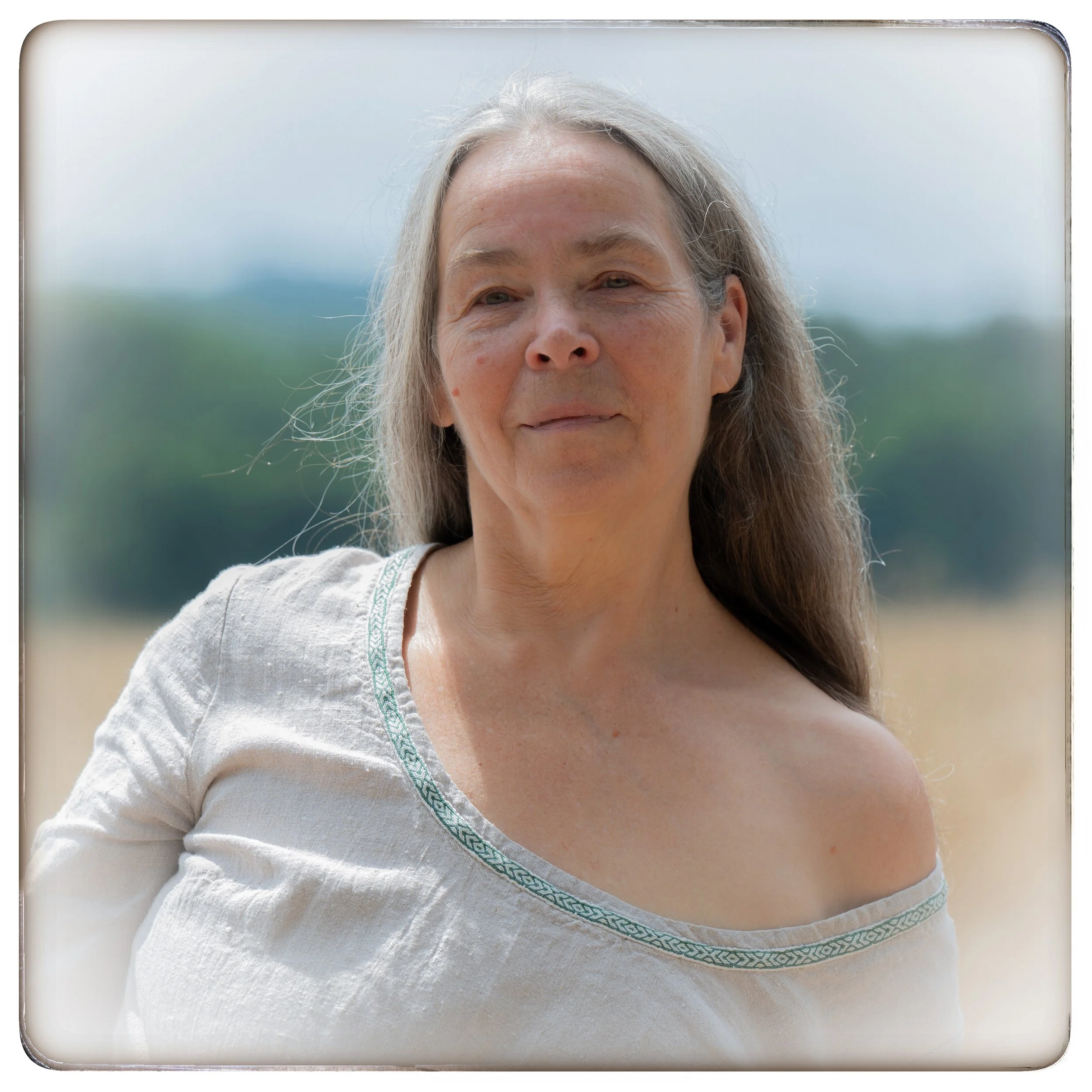 Nahaufnahme einer älteren Frau mit langem, grauem Haar, in einem weißen Top mit grüner Stickerei am Ausschnitt, im Freien mit unscharfem Landschaftshintergrund.