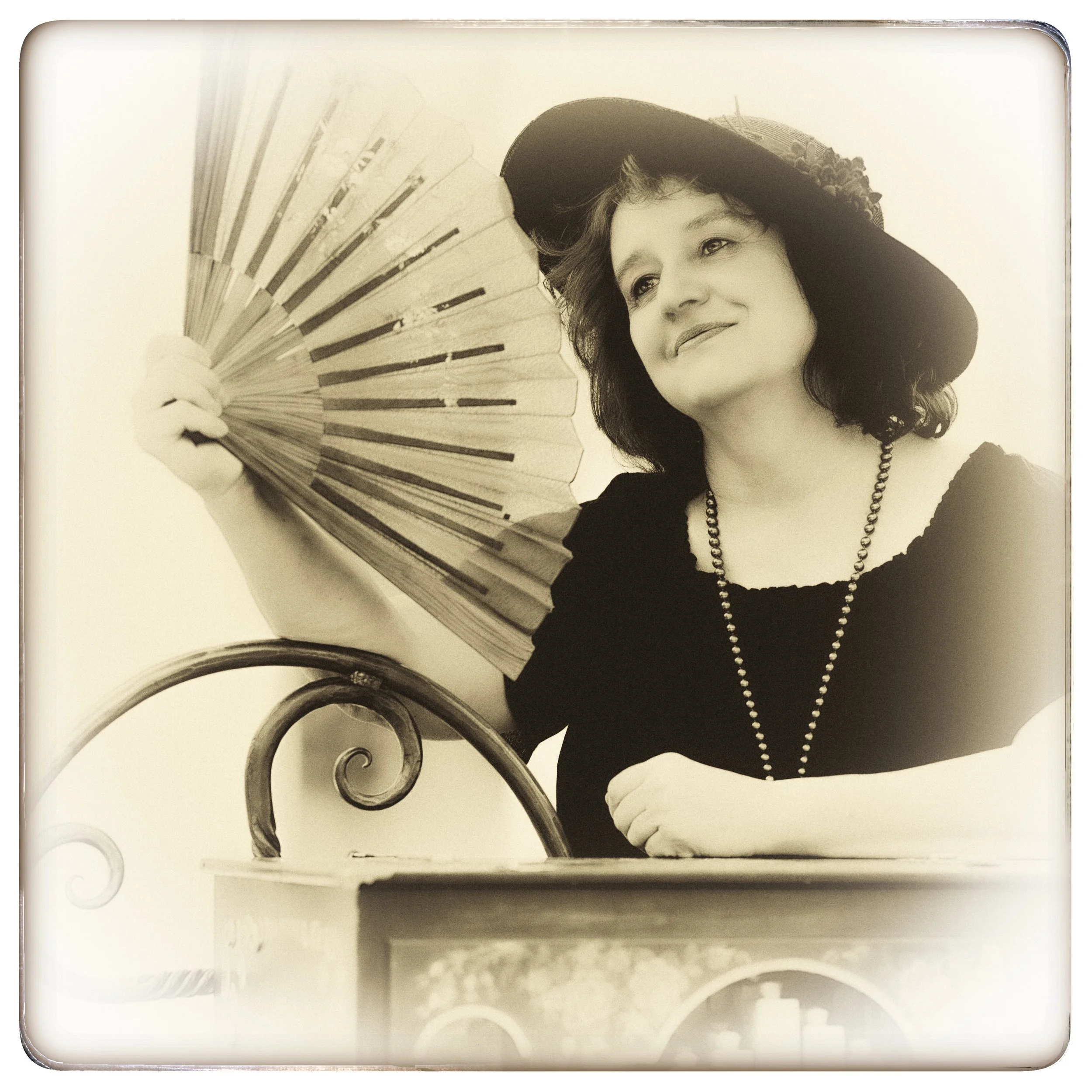 Schwarz-weiß-Foto einer Frau mit Hut, die einen Fächer hält, sitzt an einem Tisch.