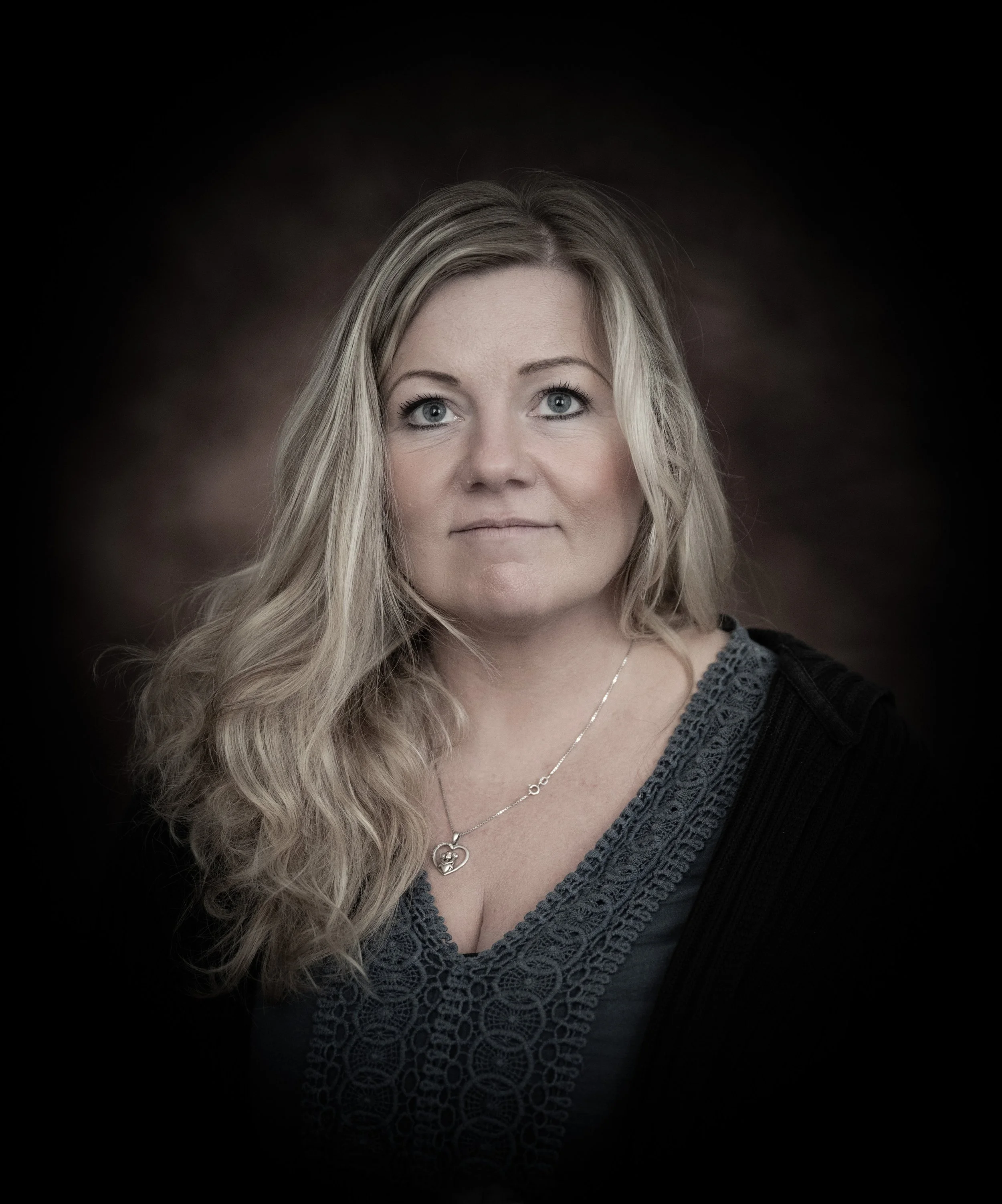 Porträt einer Frau mit blonden, gelockten Haaren und blauen Augen, trägt ein graues Top mit Spitze und eine Kette mit Herzanhänger, vor dunkelbraunem Hintergrund.