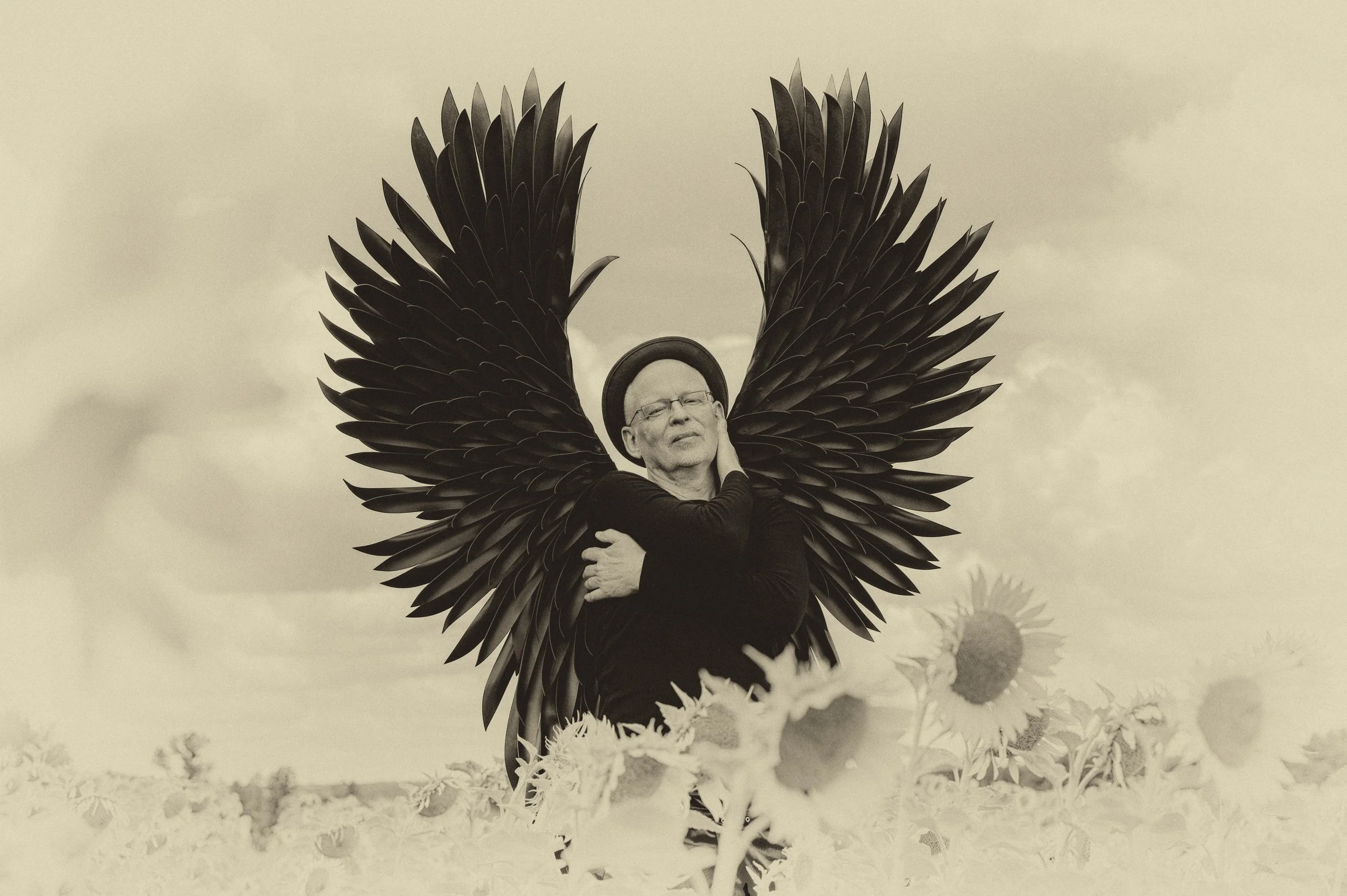 Ein älterer Mann mit Brille und Hut, der in einem Sonnenblumenfeld steht und große dunkle Flügel trägt, die wie Vogel- oder Engelsflügel aussehen.