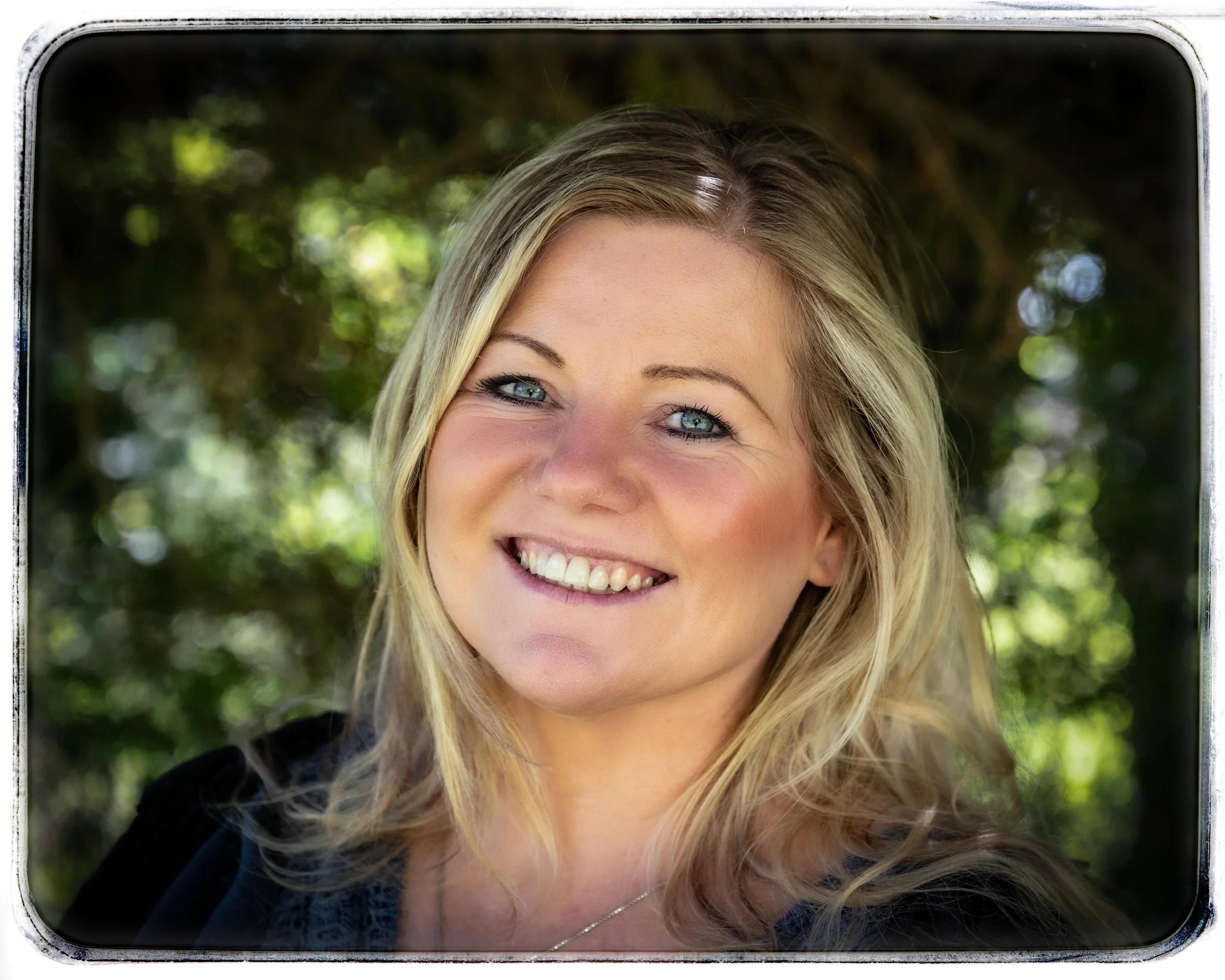 Nahaufnahme einer lächelnden jungen Frau mit blonde, lockiger Haaren vor einem grünen, bewaldeten Hintergrund.