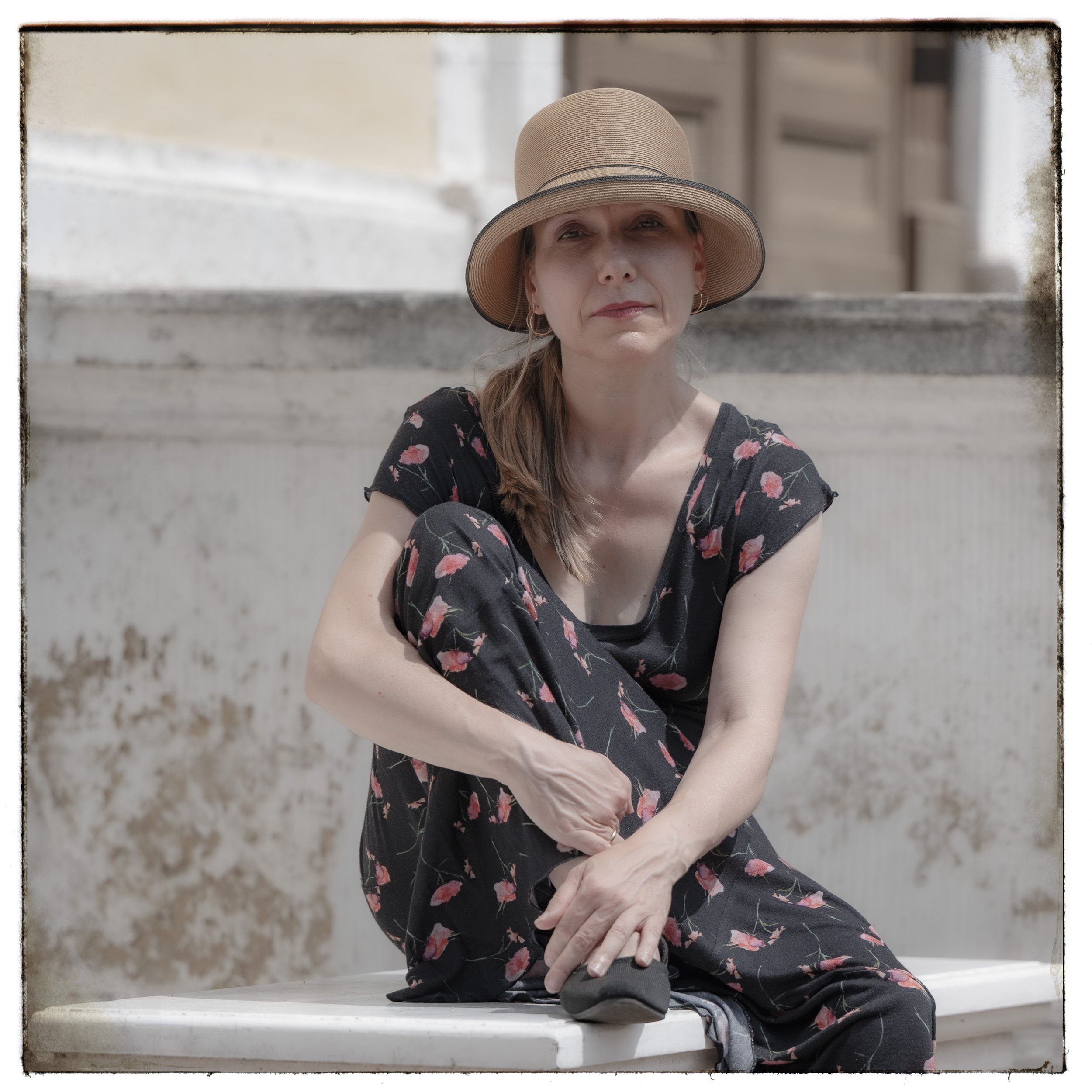 Eine ältere Frau sitzt auf einer Mauer in der Sonne, trägt einen Strohhut und eine schwarze Kleidung mit rosa Blumenmuster, hält eine Fernbedienung in der Hand, im Hintergrund ein altes Gebäude.
