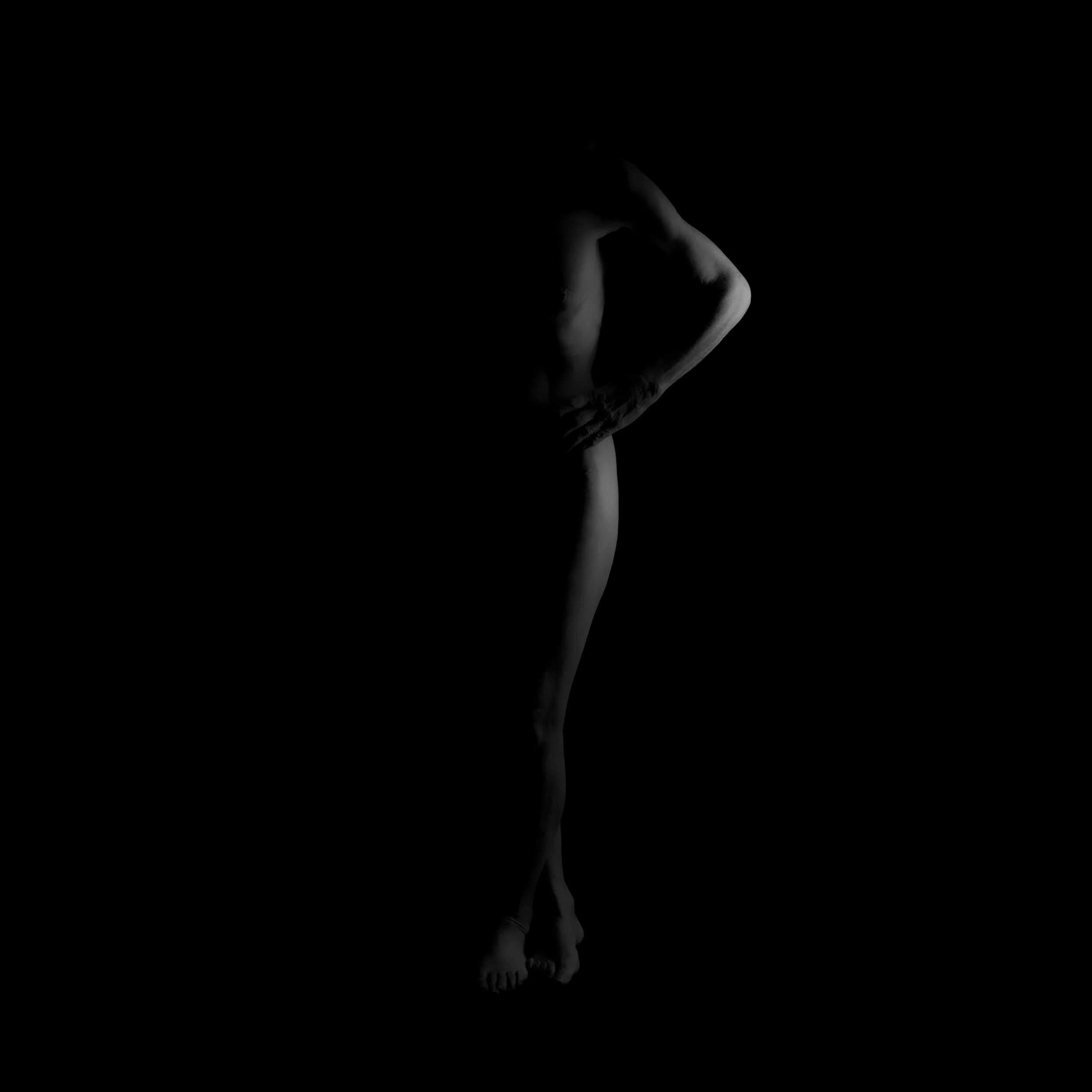 Schwarz-weiß-Fotografie einer nackten Person, die sich vor einem schwarzen Hintergrund befinde, nur teilweise sichtbar mit Beleuchtung, die die Konturen hervorhebt.