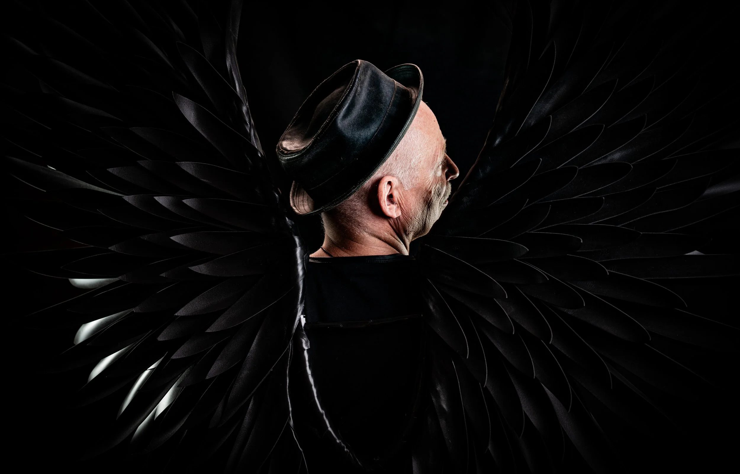 Ein älterer Mann mit weißen Haaren und einem schwarzen Hut, der in schwarze Flügel gehüllt ist.