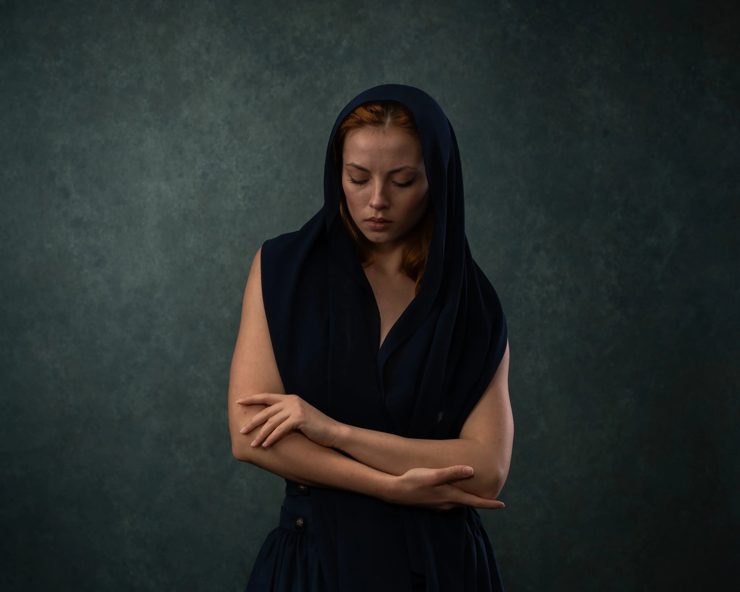 Eine junge Frau mit roten Haaren trägt ein schwarzes Kleid und einen schwarzen Schal über dem Kopf. Sie hat die Arme verschränkt und schaut nach unten.