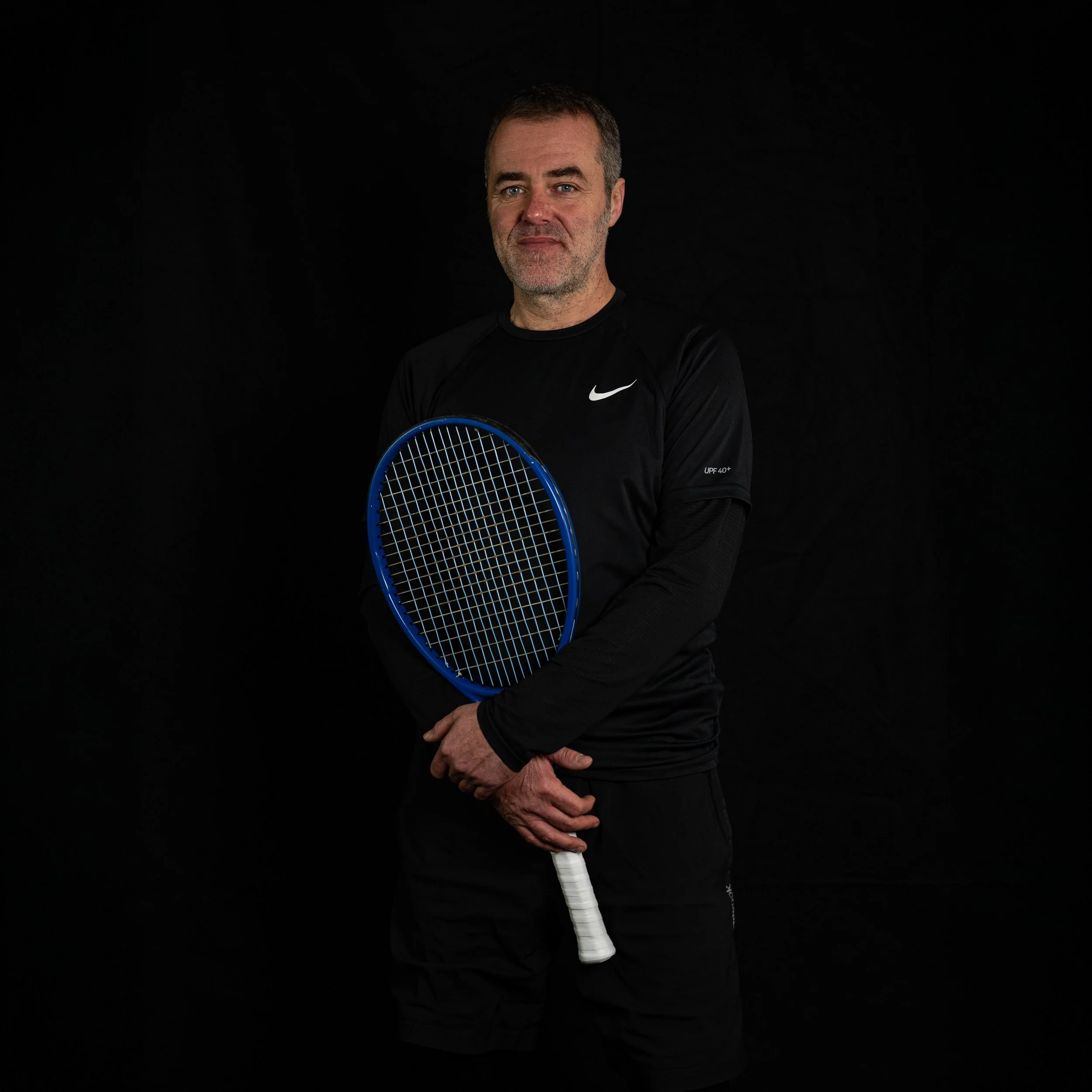 Ein Mann in schwarzer Sportbekleidung hält einen Tennis-Schläger vor einem schwarzen Hintergrund.