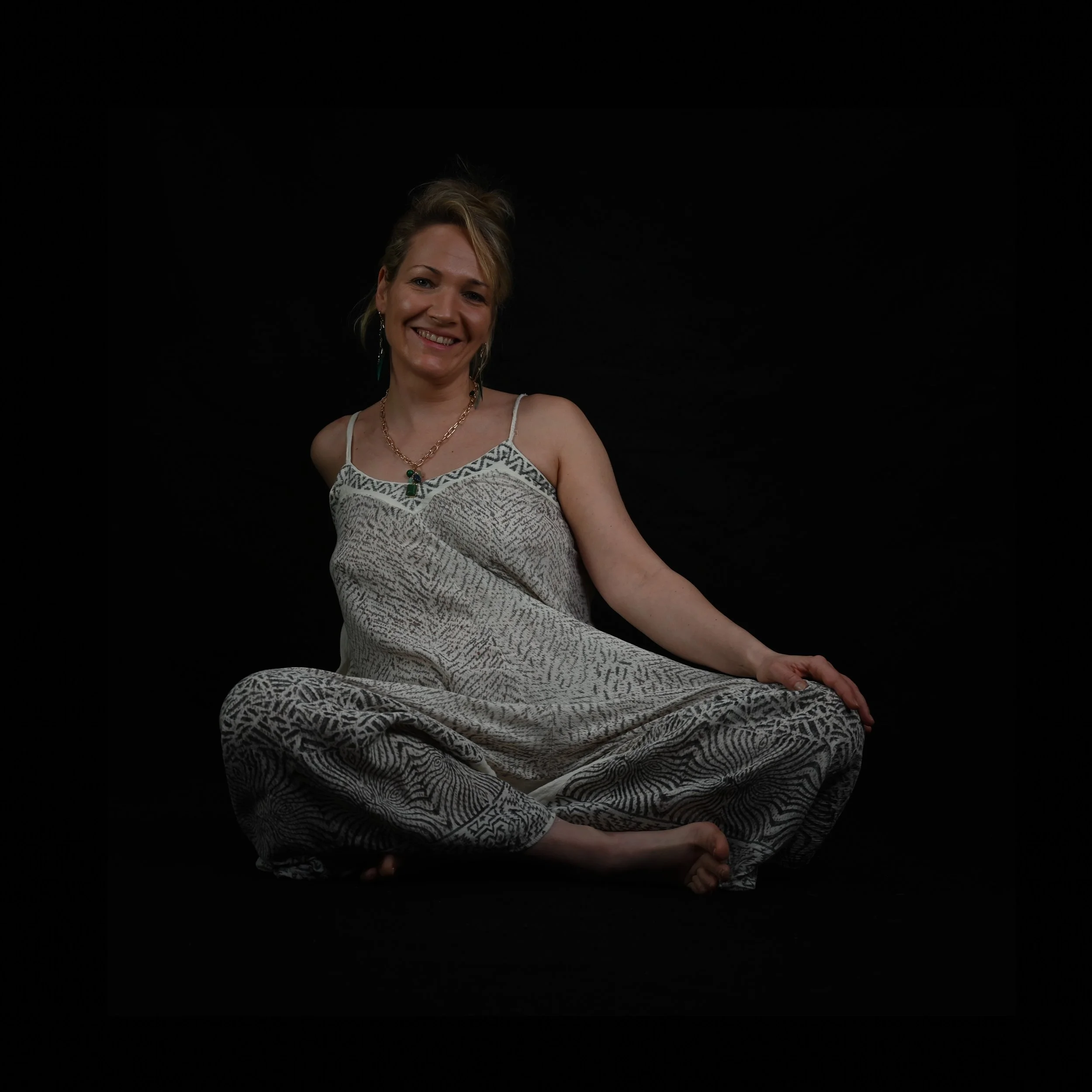 Frau sitzt auf dem Boden, lächelt, trägt viskoses, gemustertes Kleid und Schmuck, vor schwarzem Hintergrund.