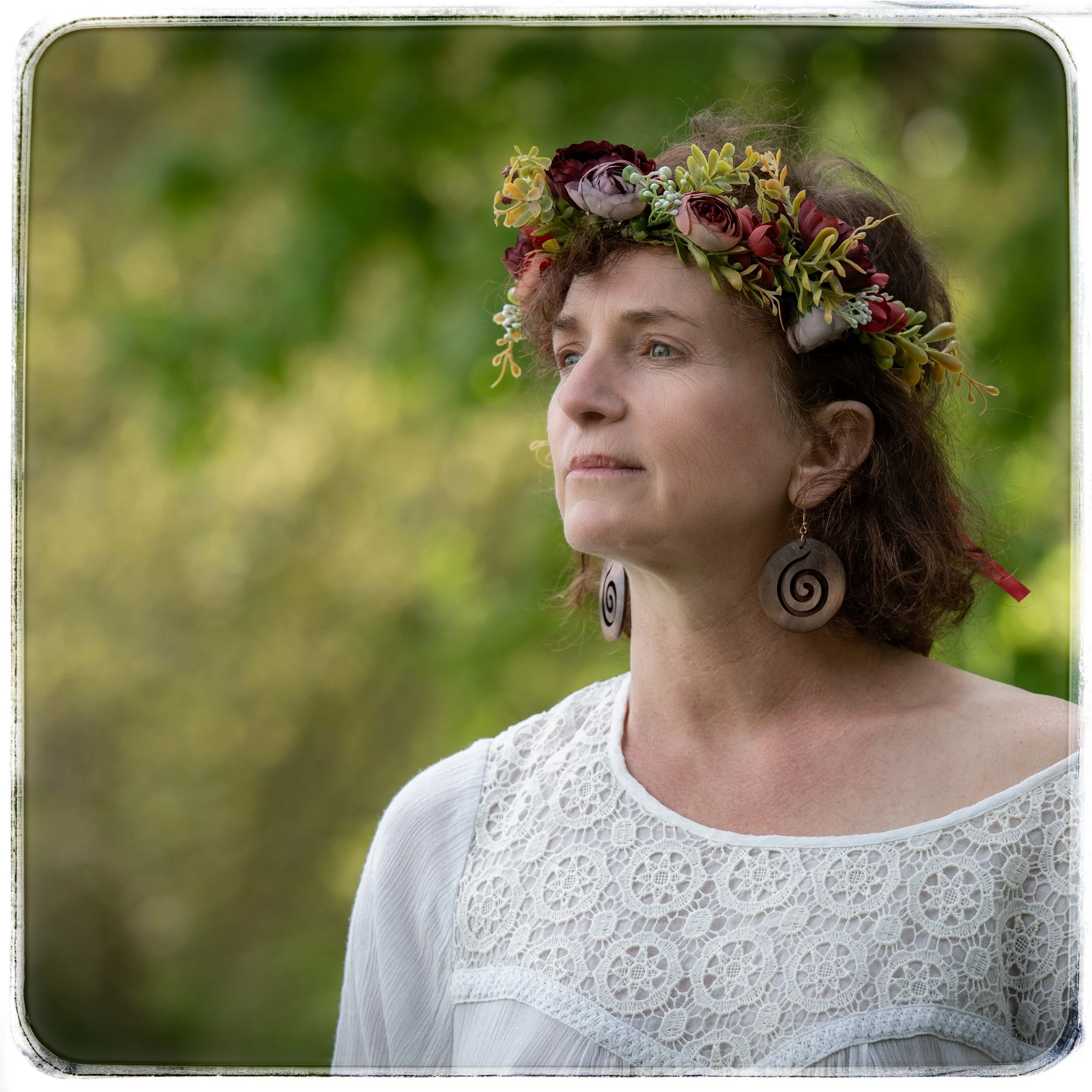 Frau mit Blumenkranz und großen spiralförmigen Ohrringen, trägt ein weißes Spitzenkleid, im Freien mit grüner Baumkulisse