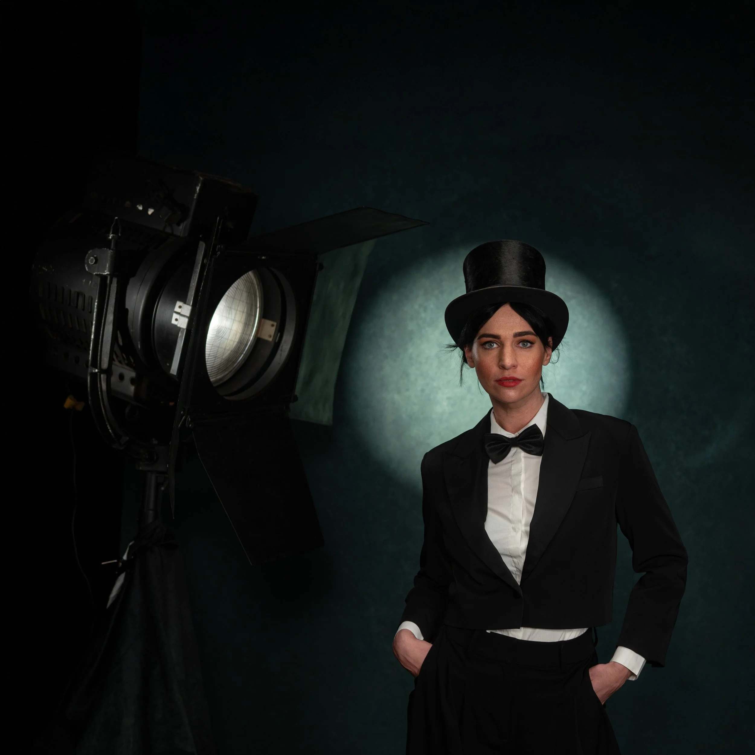 Frau in formellem schwarzen Anzug mit Hut und Fliege vor dunklem Hintergrund, Beleuchtung durch Studio-Licht.