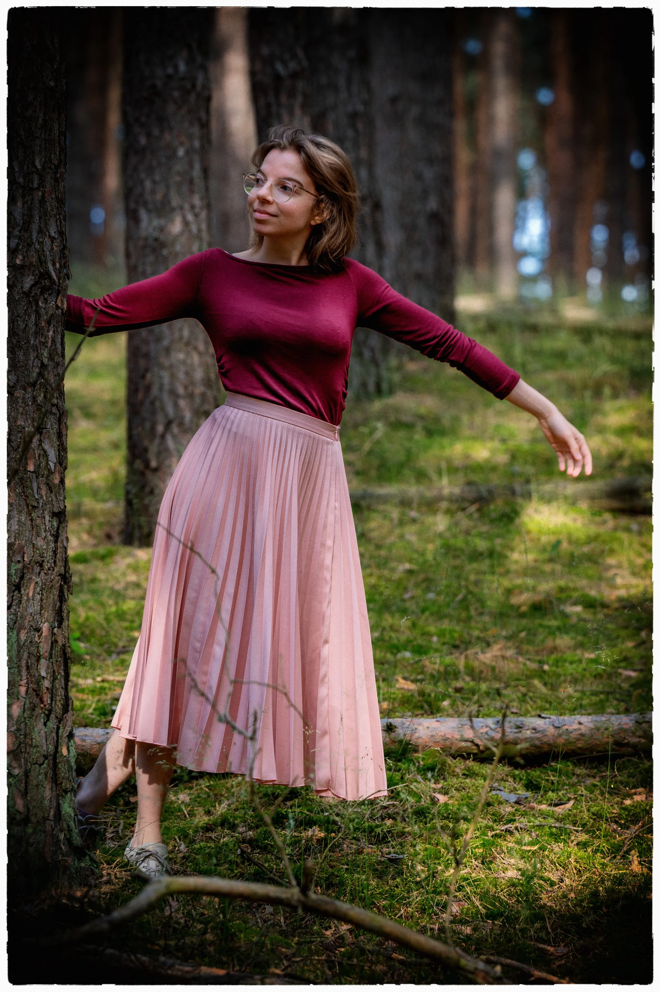 Frau steht in einem Wald, trägt einen burgunderroten Pullover und eine hellrosa, plissierte Röcke, sieht entspannt und poised aus.