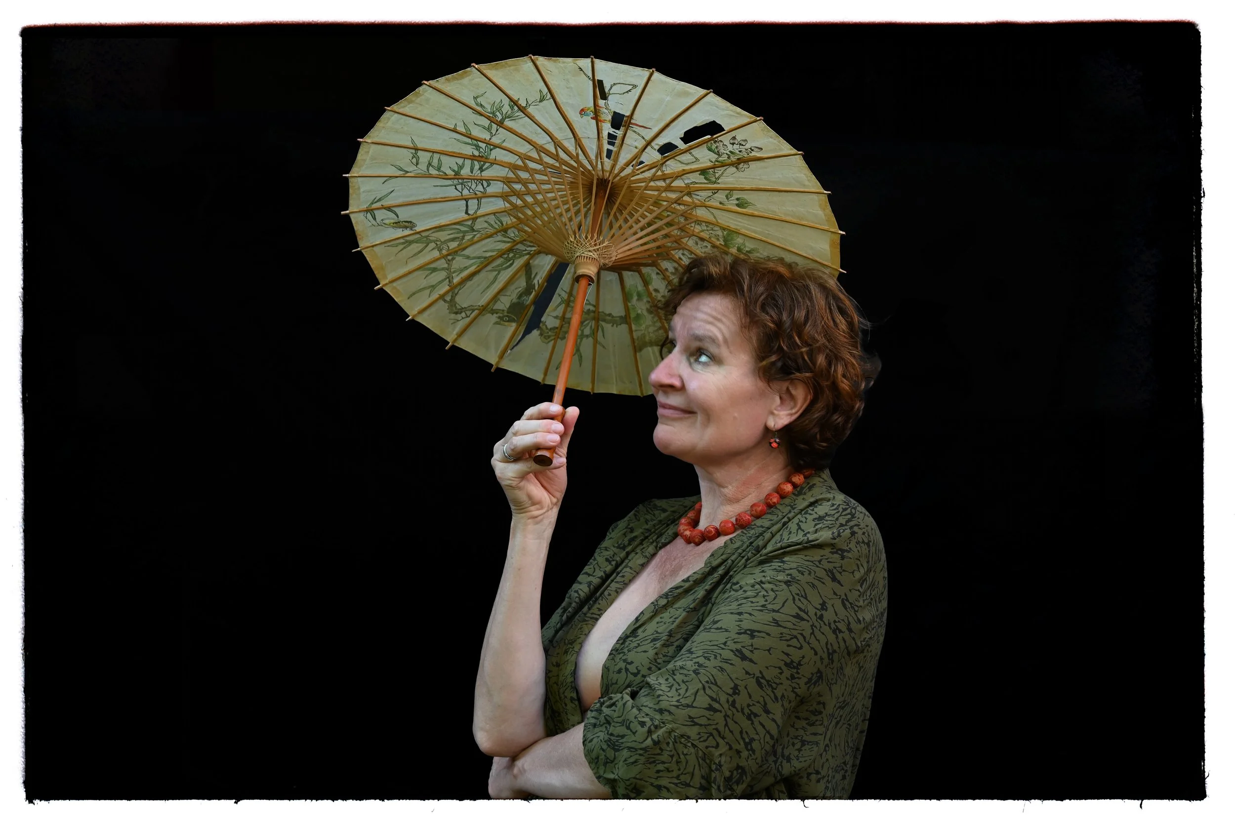 Eine Frau mit kurzen, lockigen braunen Haaren hält einen traditionellen japanischen Papierschirm, schaut nach oben und lächelt. Sie trägt eine grüne Jacke mit Muster und eine rote Halskette vor schwarzem Hintergrund.