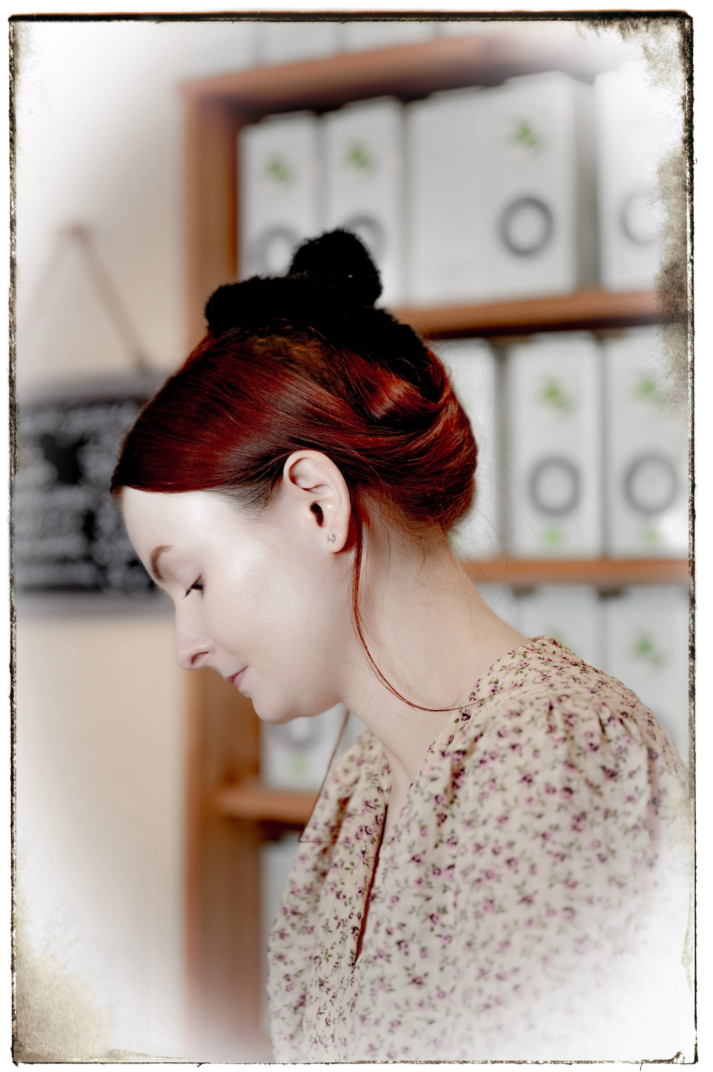 Eine junge Frau mit roten Haaren, seitlich fotografiert, trägt einen schwarzen Haarschmuck und ein blumiges Oberteil, im Hintergrund sind Bücherregale mit weißen Ordnern sichtbar.