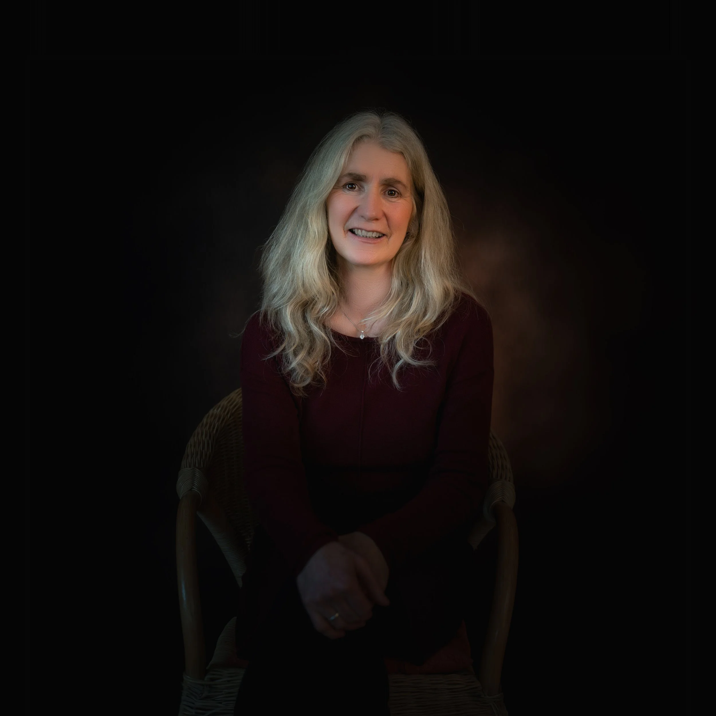 Porträt einer lächelnden Frau mit langen, lockigen blondem Haar, sitzend auf einem Stuhl gegen dunklem Hintergrund.