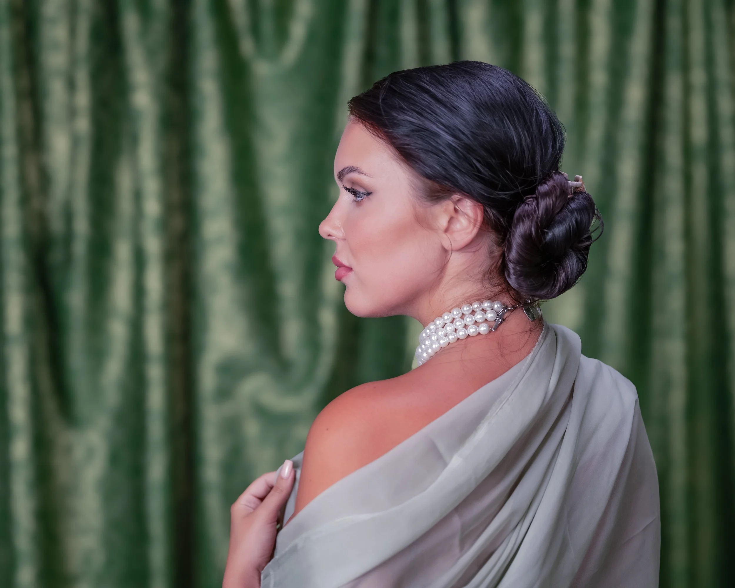 Profilaufnahme einer eleganten Frau mit dunklem, hochgestecktem Haar, Perlenkette, Schulterkleid vor grünem, seidigem Hintergrund.