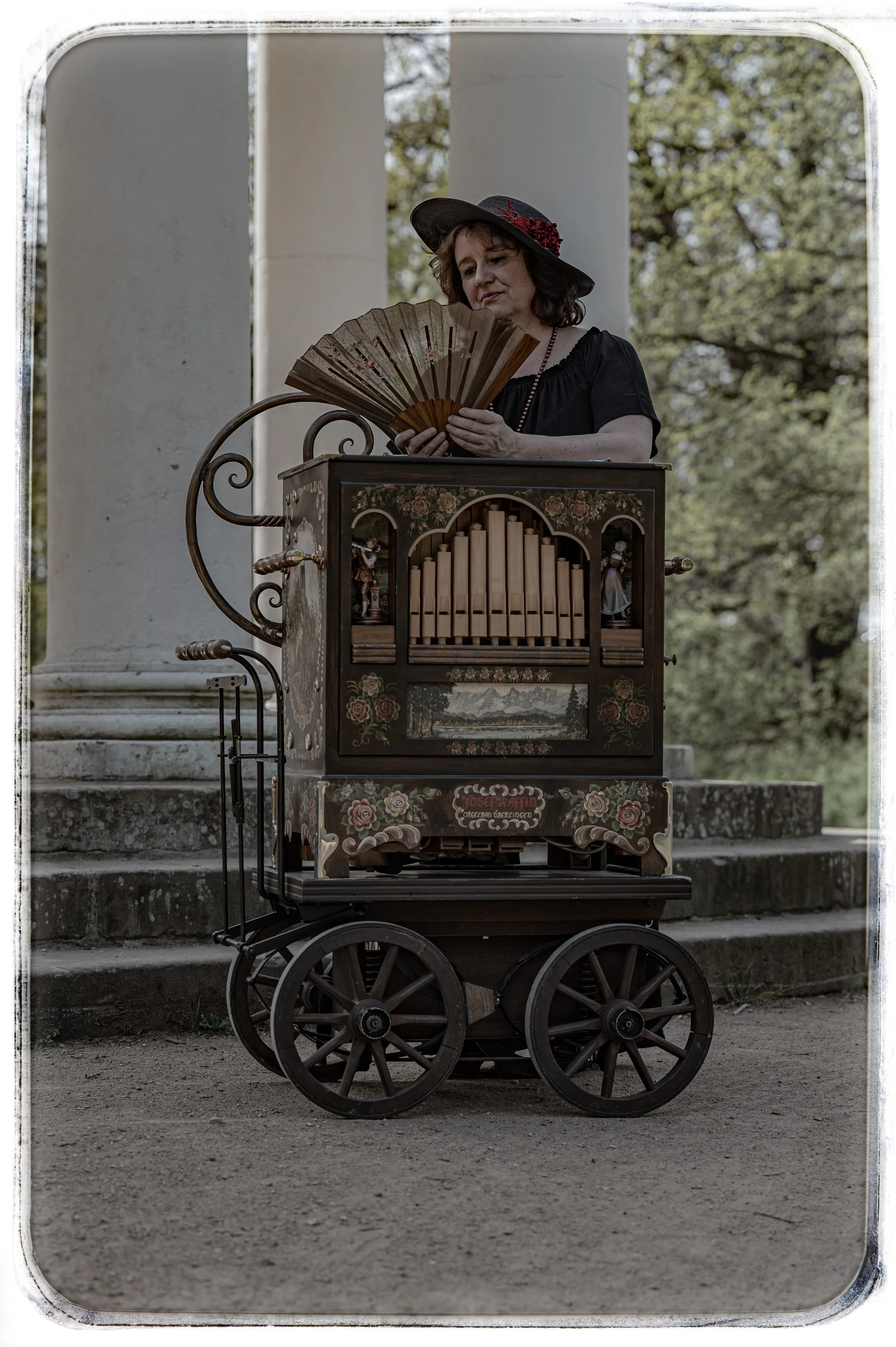 Frau in historische Kleidung mit Hut spielt eine handbetriebene Orgeldarstellung im Freien.