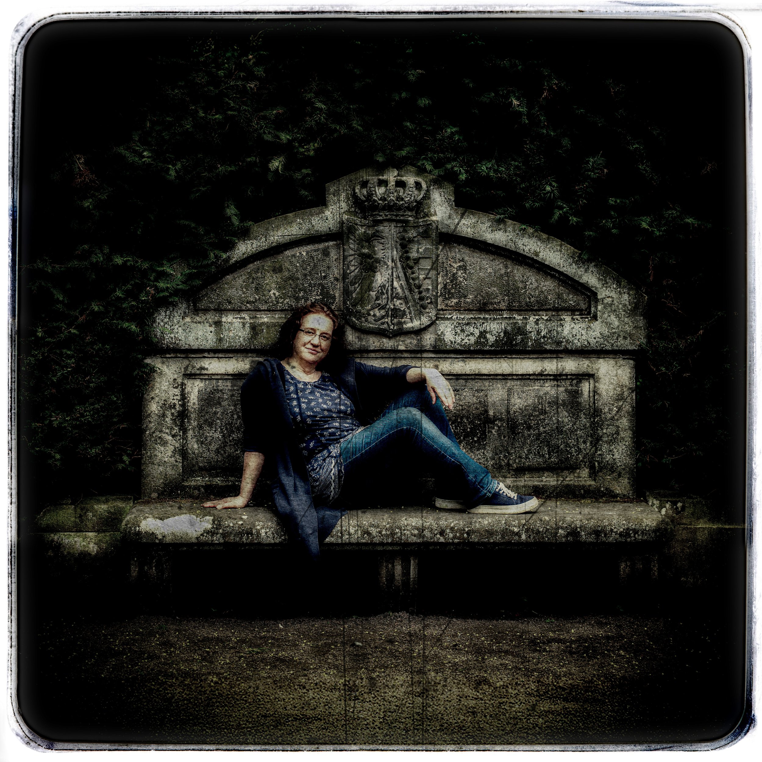 Eine Frau sitzt auf einer Steinbank vor einer antiken Steinstatue mit Wappenschild, umgeben von grünen Büschen.