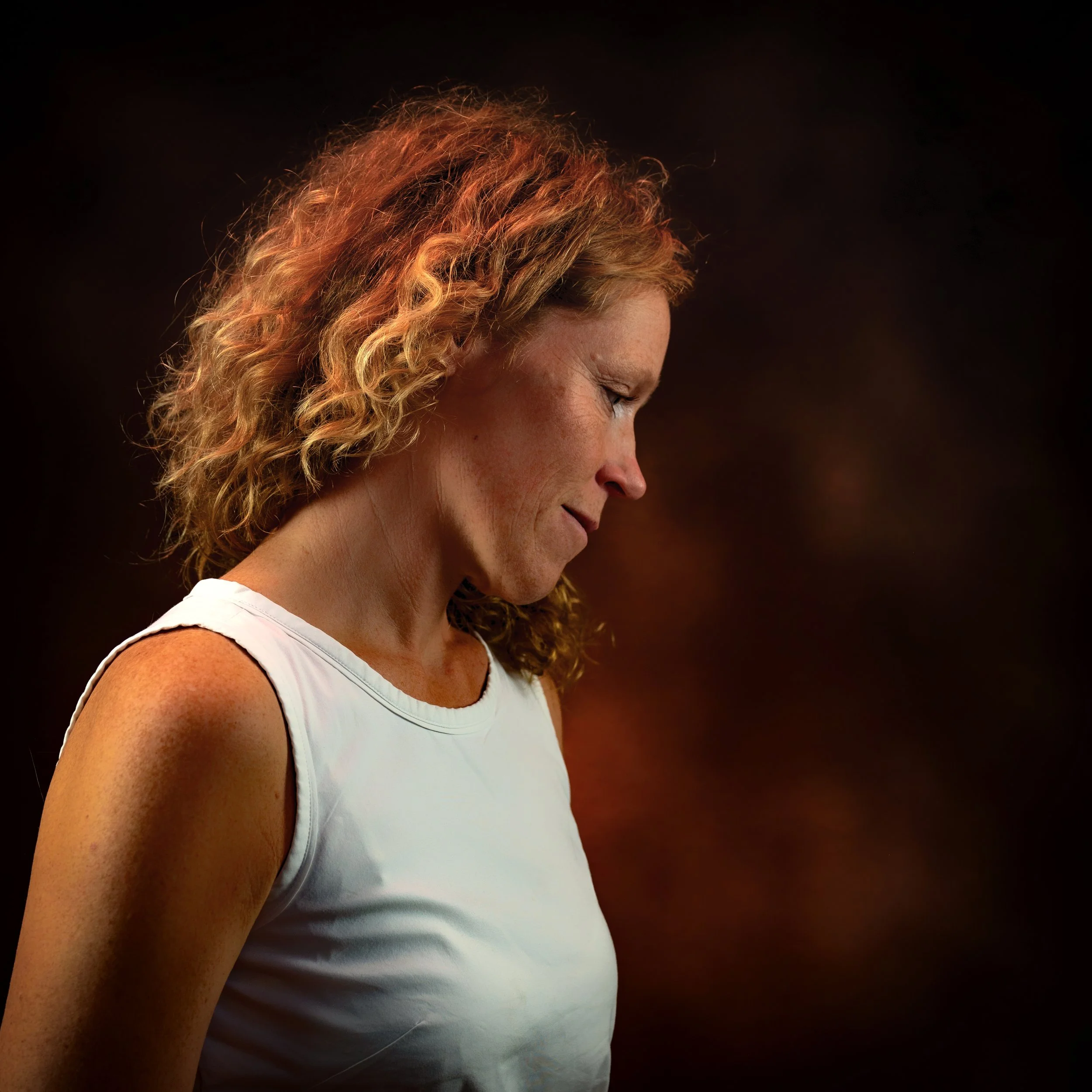 Frau mit roten, lockigen Haaren in Seitenansicht trägt weißes Top vor dunklem Hintergrund.