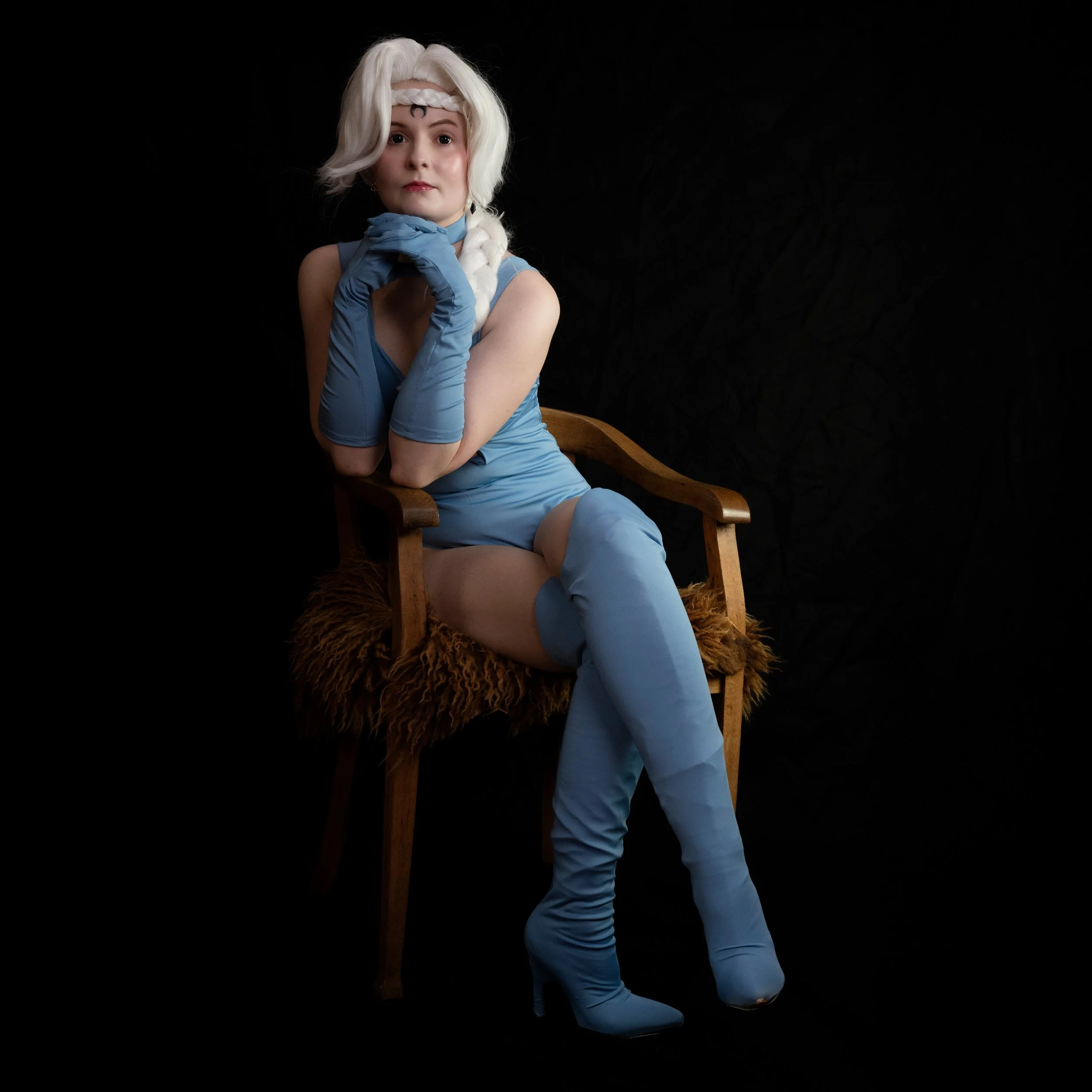 Frau in blauer Kostüm mit weißen Haaren, sitzend auf einem Holzstuhl mit Felldecke, schwarze Hintergrund.