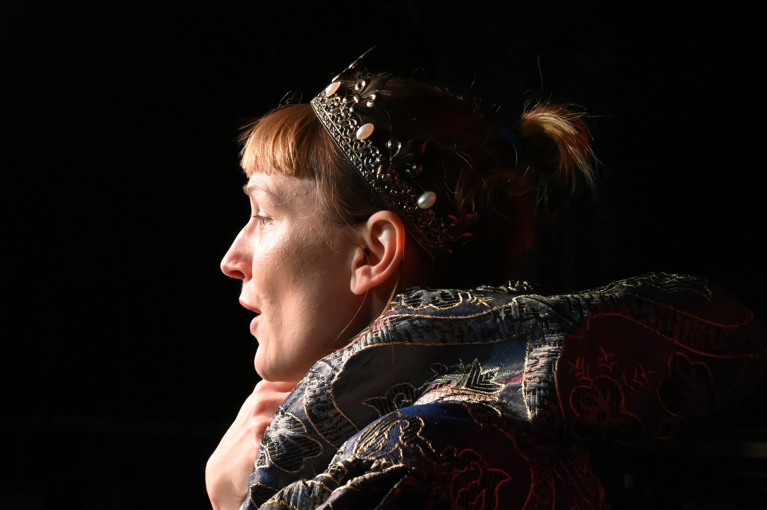 Porträt einer Frau mit rotem Haar, eine Tiara und einer schwarzen, bedruckten Jacke, sitzt im Dunkeln.